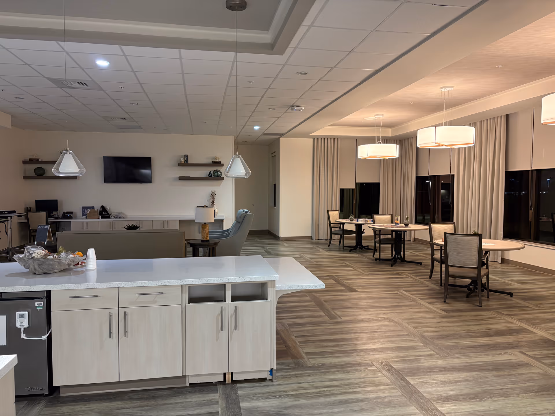 Open common area with kitchenette island, seating tables, chairs, and wall-mounted TV.