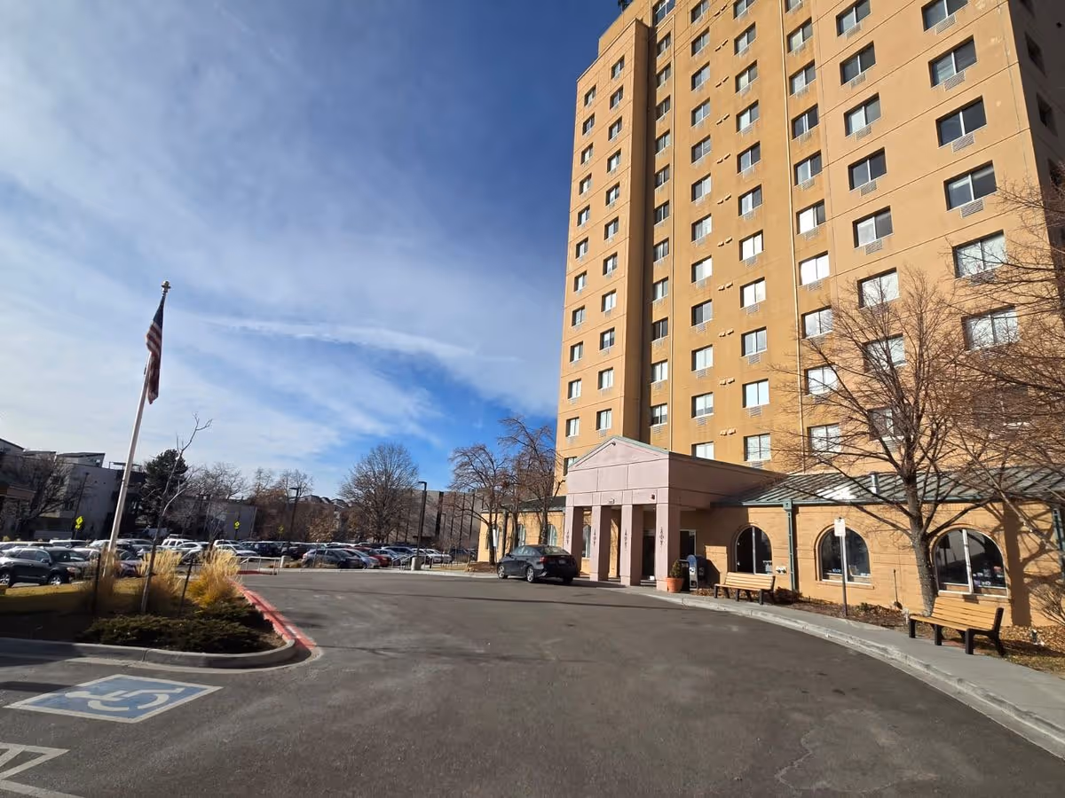 Multi-story senior living building entrance with a circular driveway, benches, parked cars, and an American flag under a blue sky.