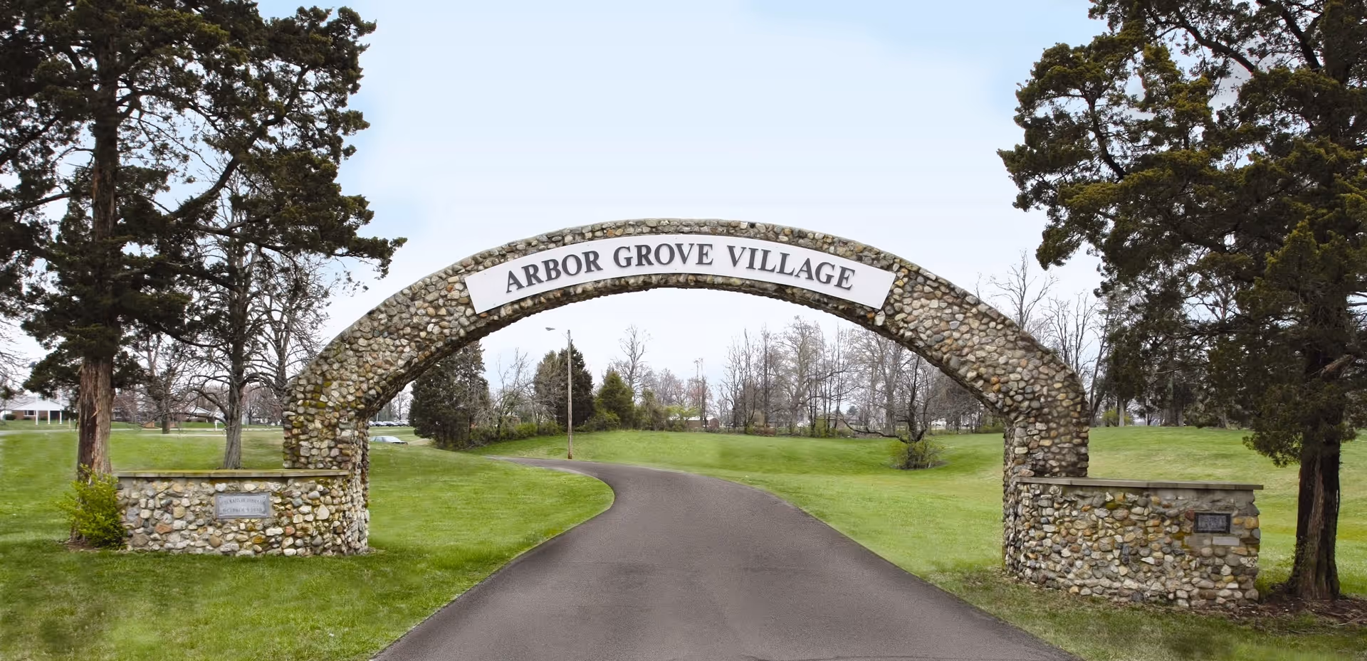 Stone arch entrance reading 'Arbor Grove Village' over a paved driveway surrounded by grassy lawns and trees.