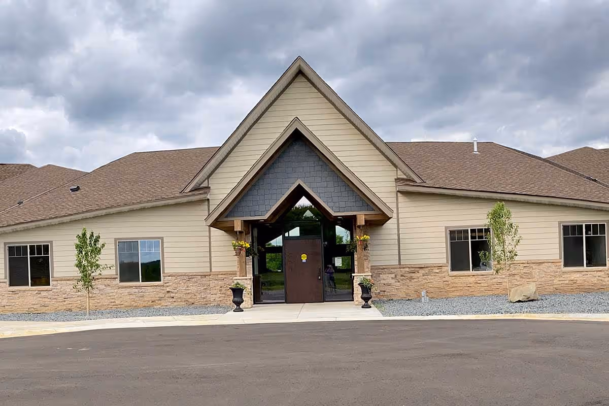 Front exterior view of a single-story building with beige siding, stone accents, a brown shingled roof, and a prominent triangular entrance with glass doors. There are small trees and planters with flowers near the entrance under a cloudy sky.