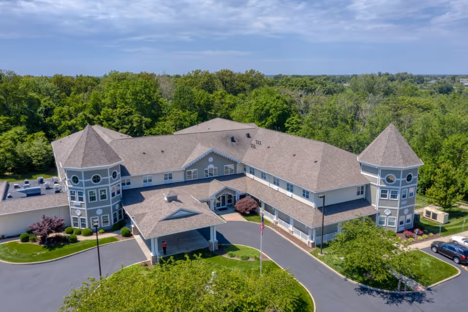 Aerial view of a large senior living facility building with two prominent turret-style towers on each end, surrounded by green trees and landscaping. The building has a covered entrance driveway and a parking area with several cars visible.
