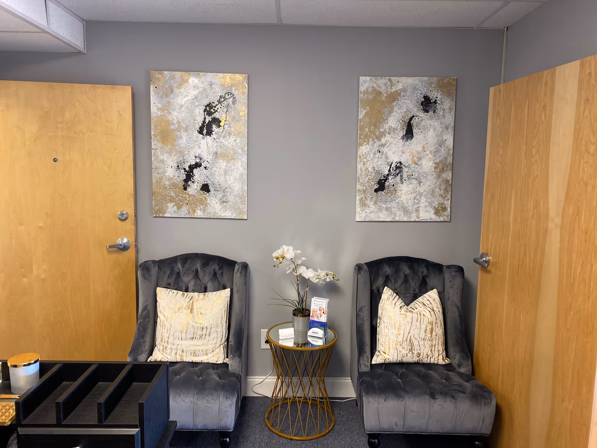 Two tufted gray chairs flank a small round table with flowers in a waiting area.