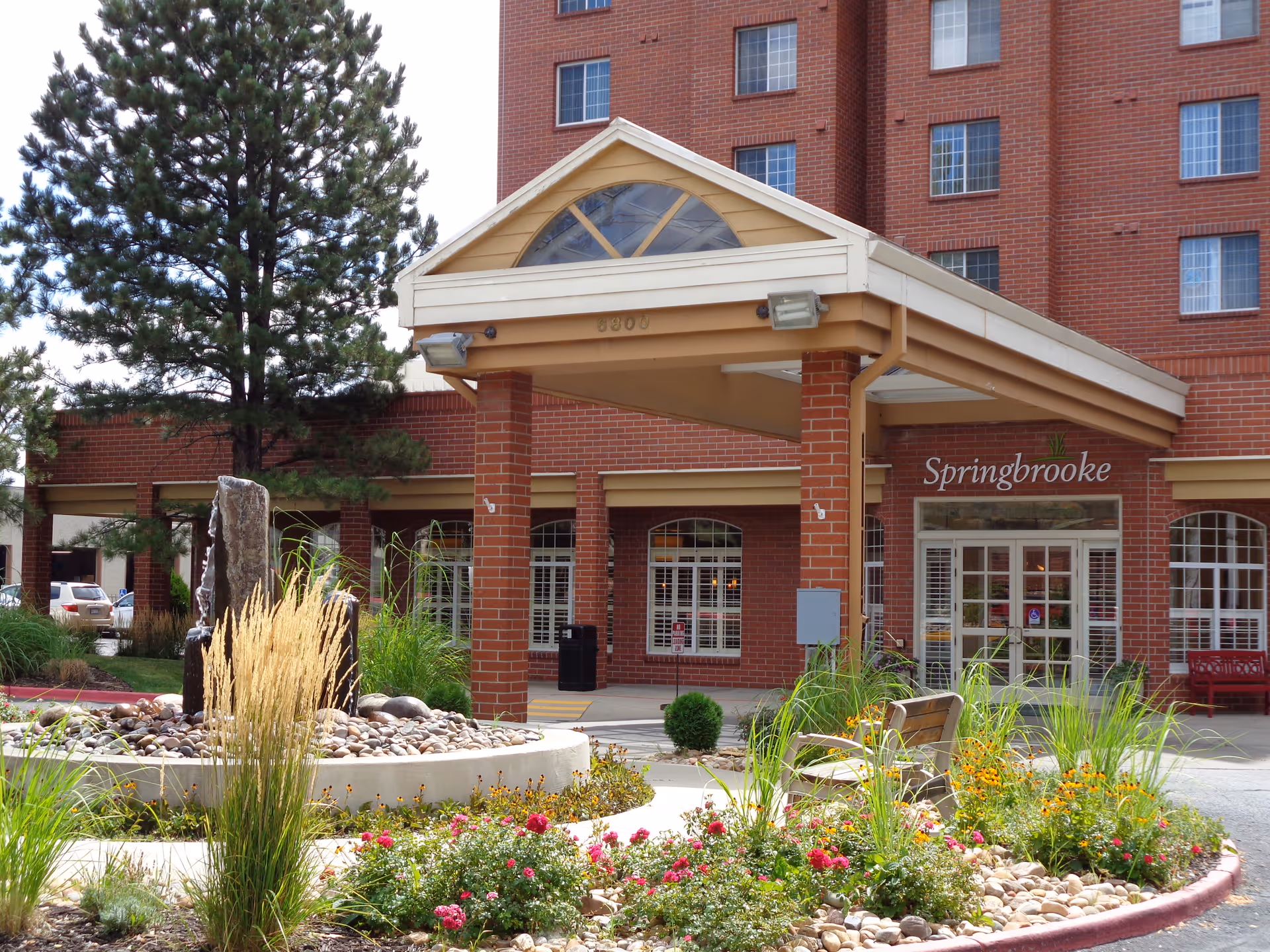 Entrance of Springbrooke Retirement facility showing a covered drop-off area with brick pillars, a landscaped garden with flowers, grasses, and rocks in the foreground, and the multi-story brick building in the background.