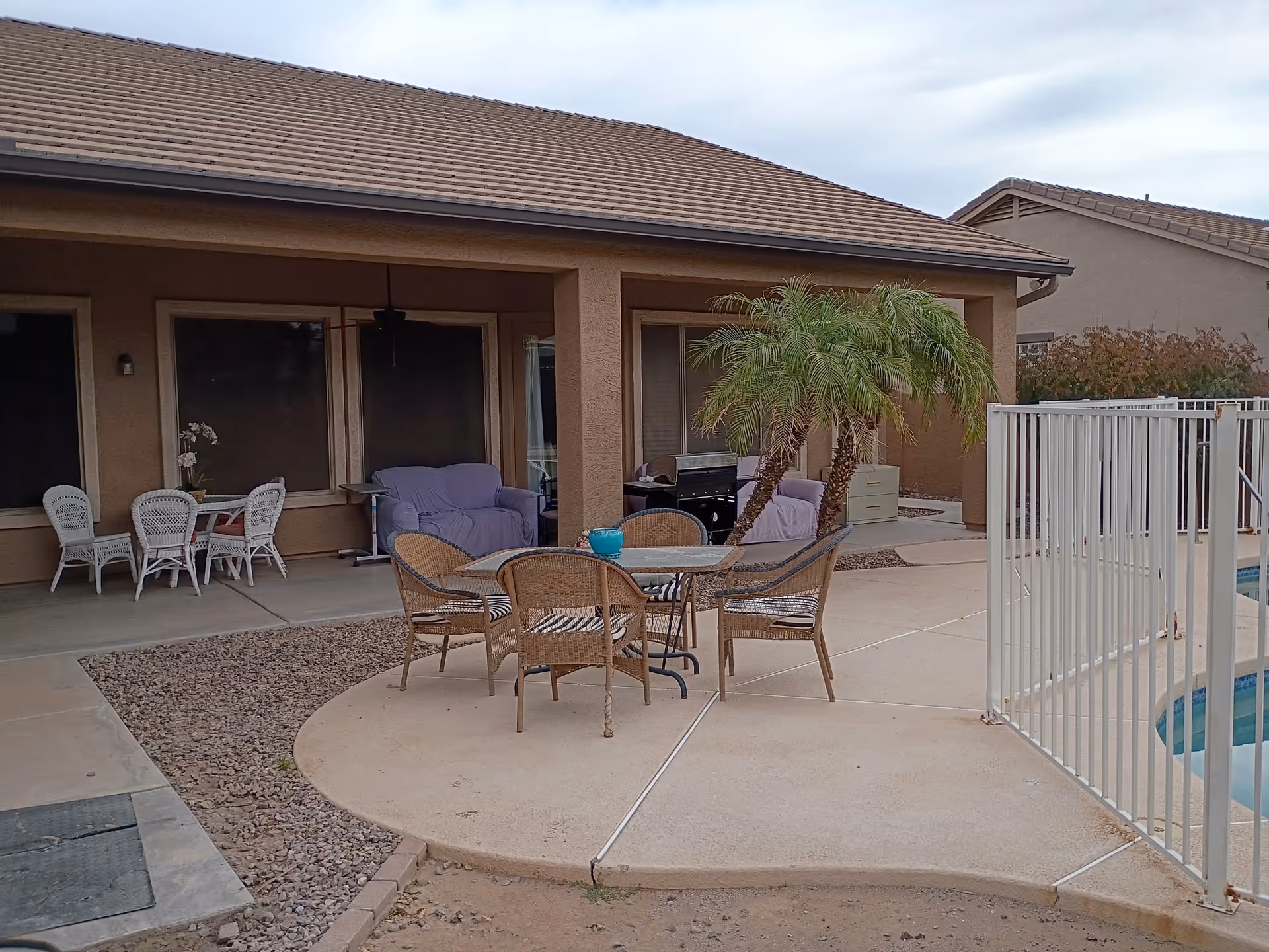 Outdoor patio with wicker furniture, palm trees, and a fenced pool behind a tan building.