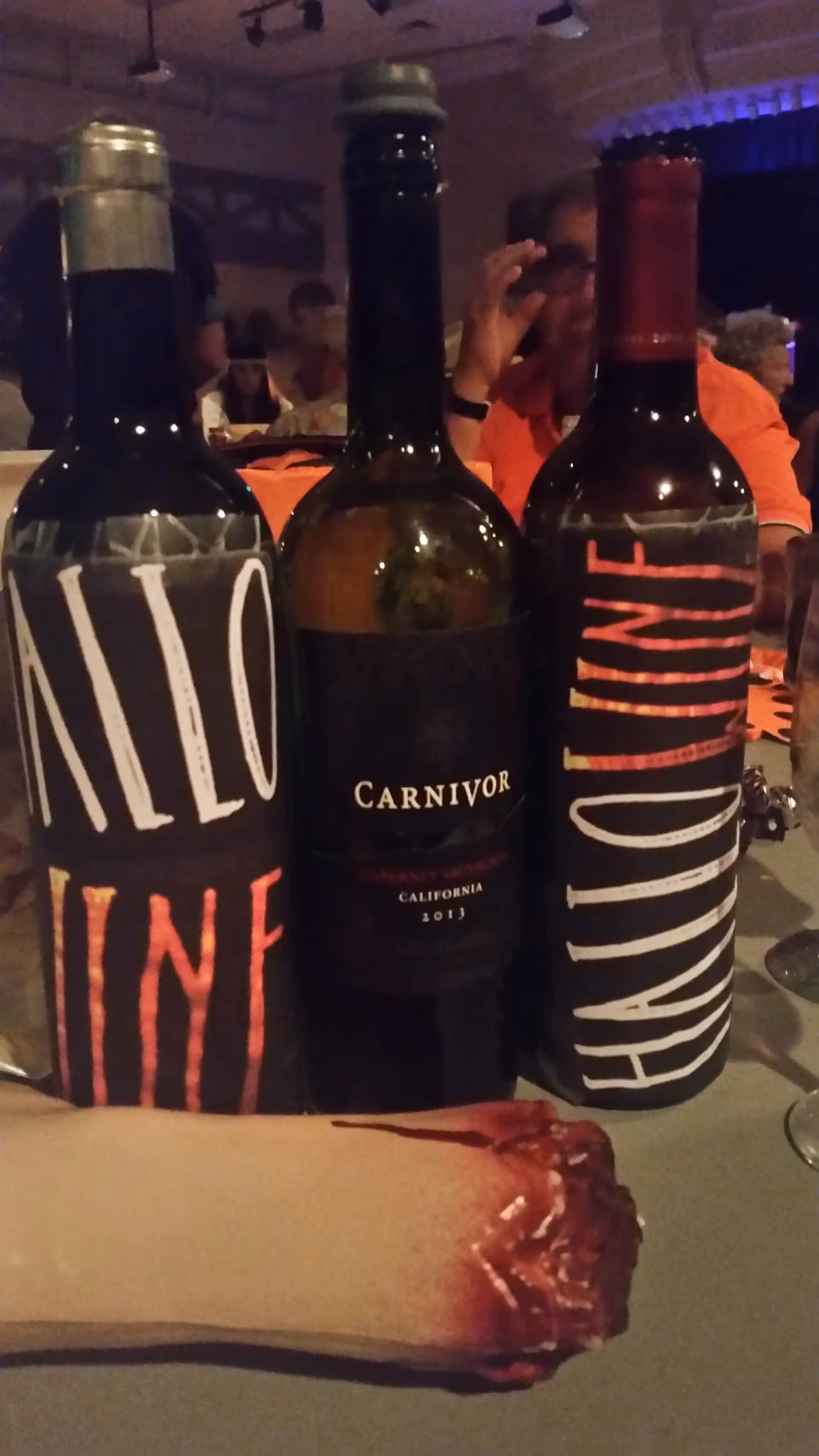 Three wine bottles stand on a dining table as diners eat in a dimly lit room.