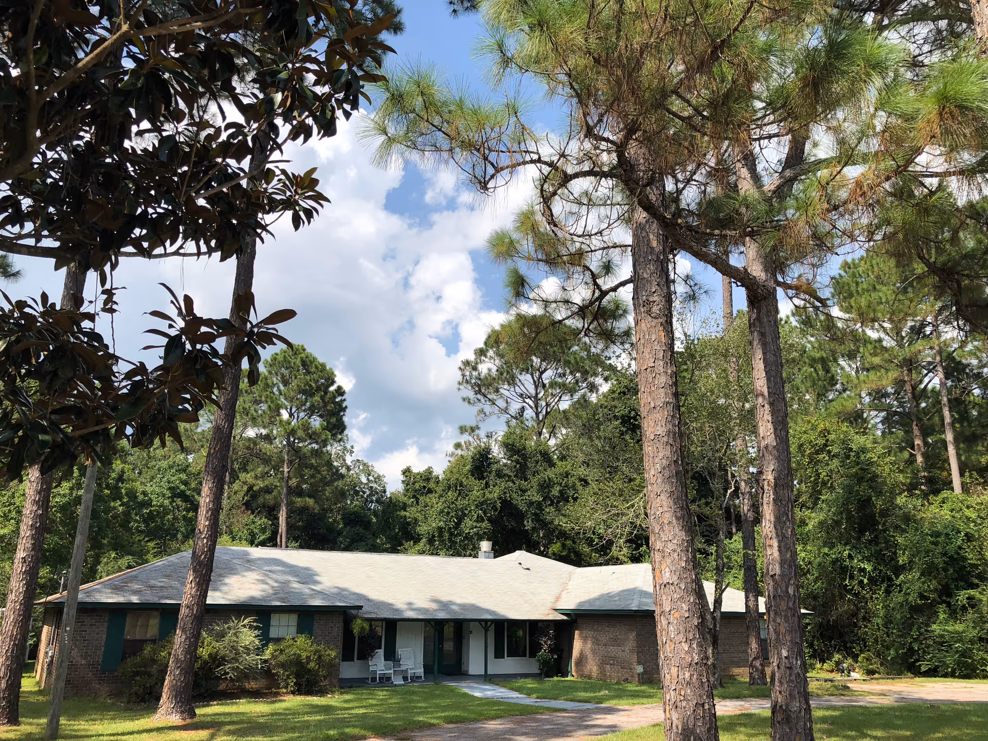 Single-story brick building with a light roof and front porch surrounded by tall pine trees and a grassy lawn under a partly cloudy sky.
