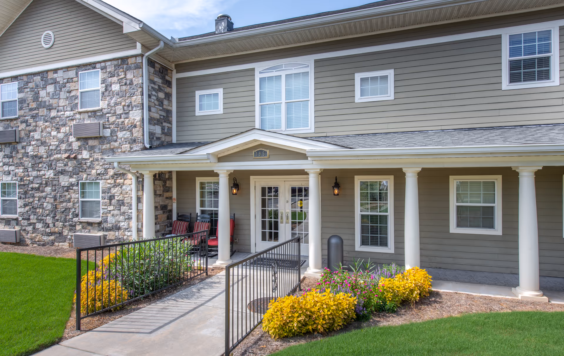 Front entrance of a senior living building with a covered porch, columns, an accessibility ramp, and landscaped flower beds.