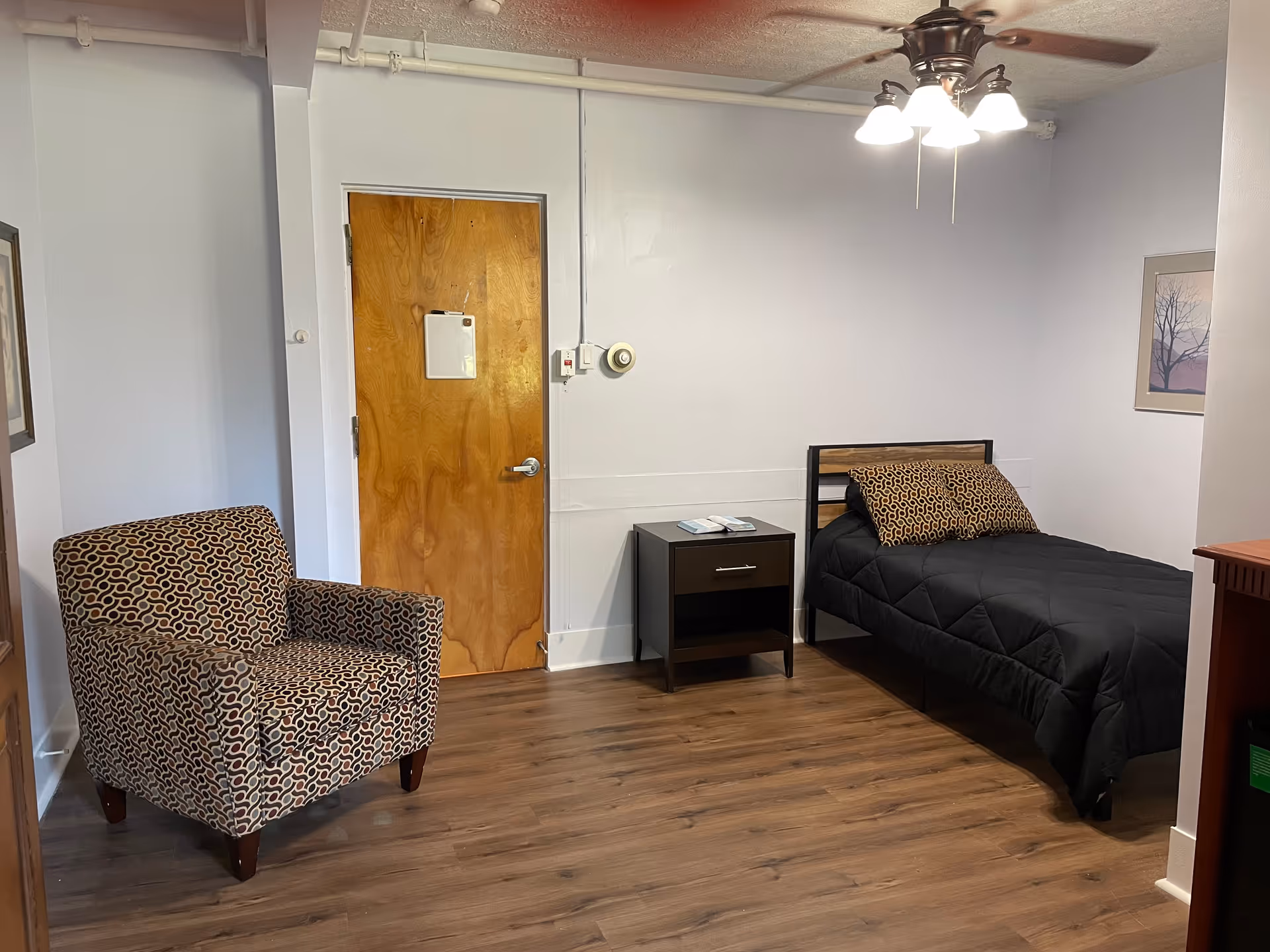 Small bedroom with a bed, nightstand, patterned chair, ceiling fan, and wooden floor.