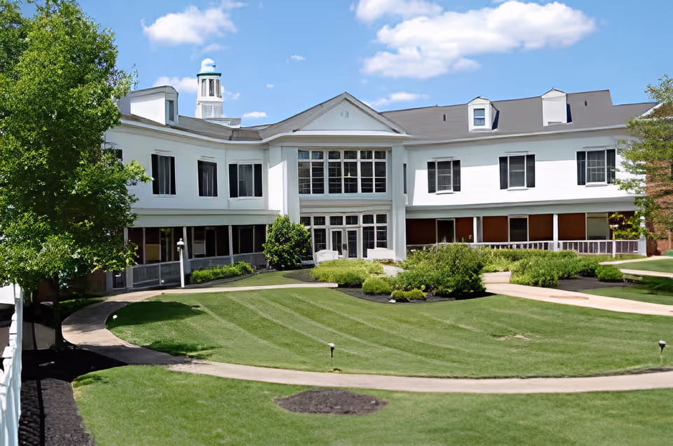 Front exterior of a white two-story assisted living building with manicured lawn, walkways, and trees under a blue sky.