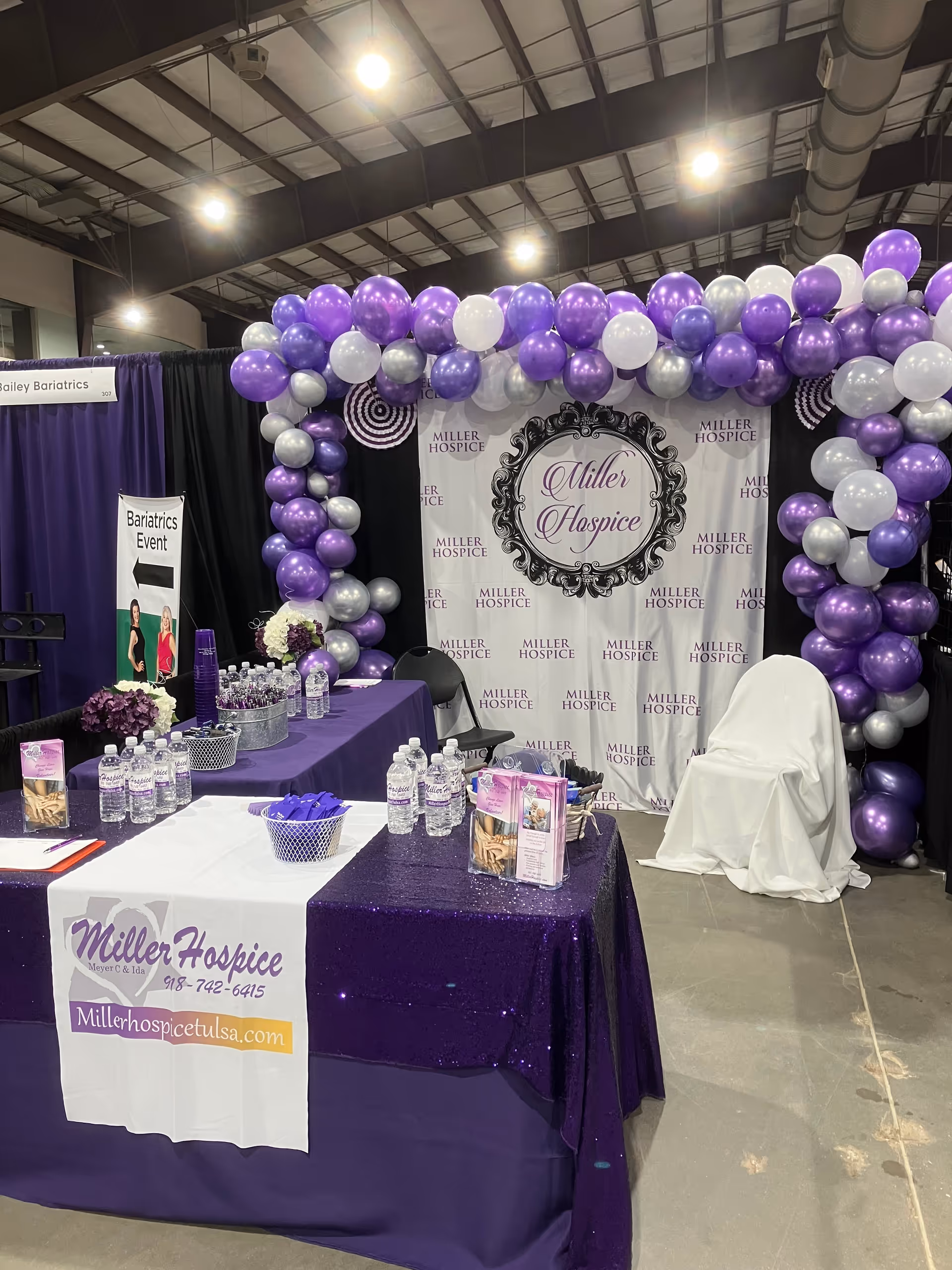 Indoor expo booth with purple balloons, Miller Hospice banners, and display tables.