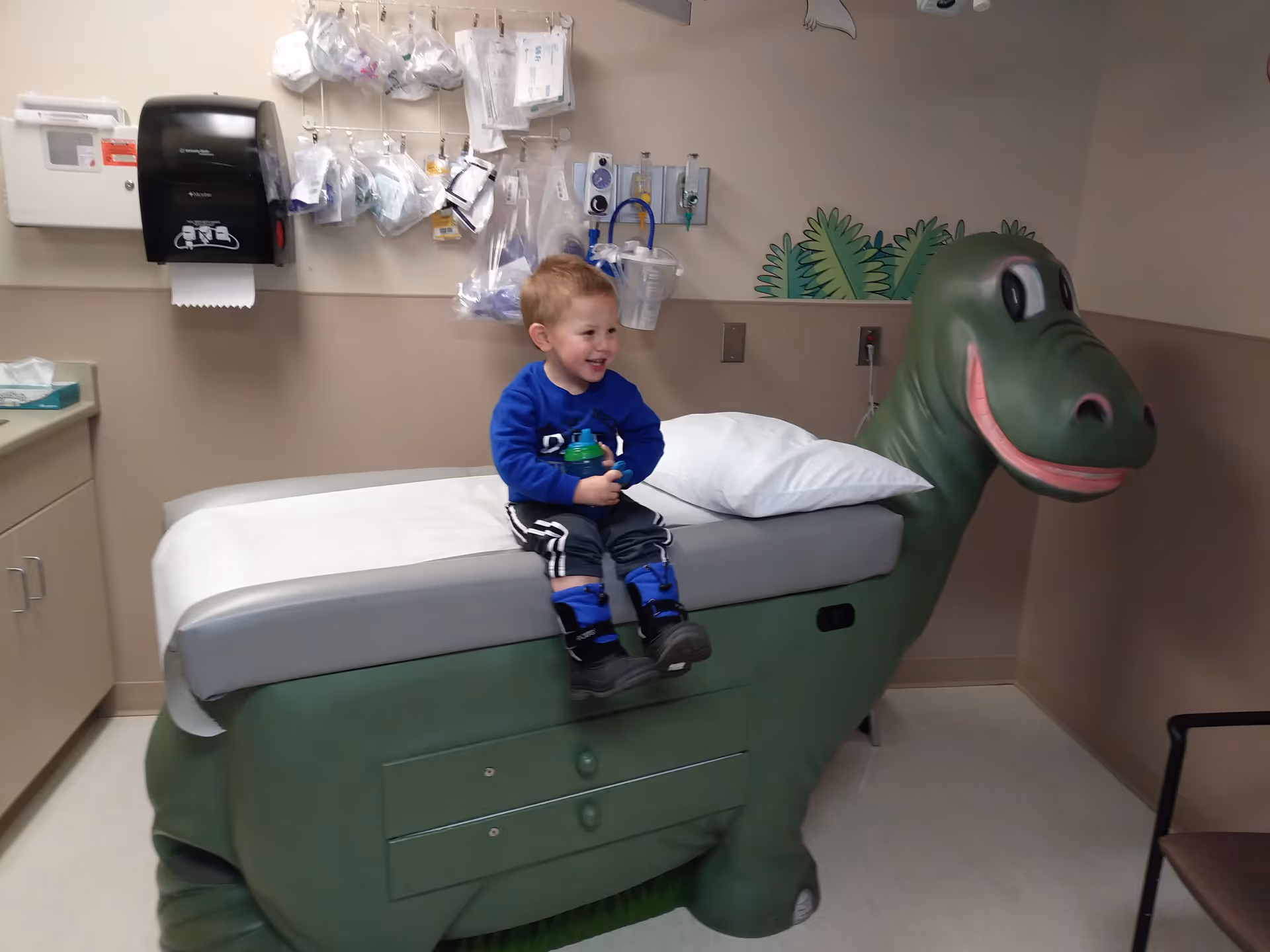 Young child sits on a dinosaur-shaped hospital bed in a clinical room.
