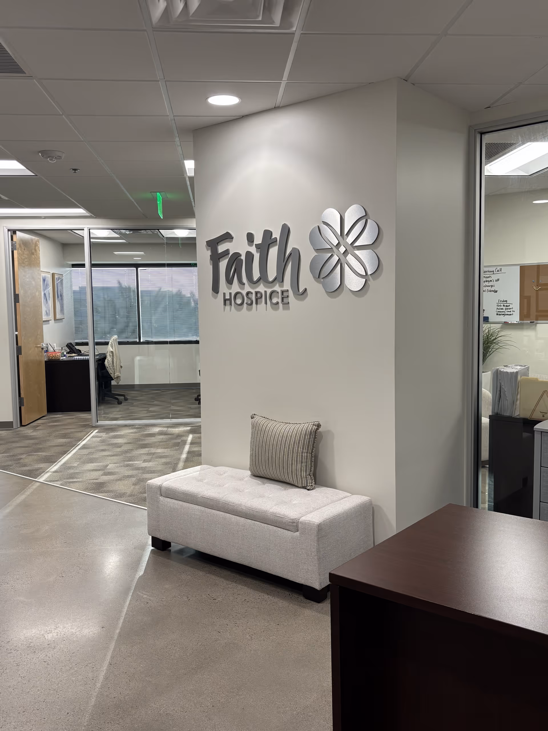 Lobby area with Faith Hospice sign, bench seating, and office workspace behind glass.