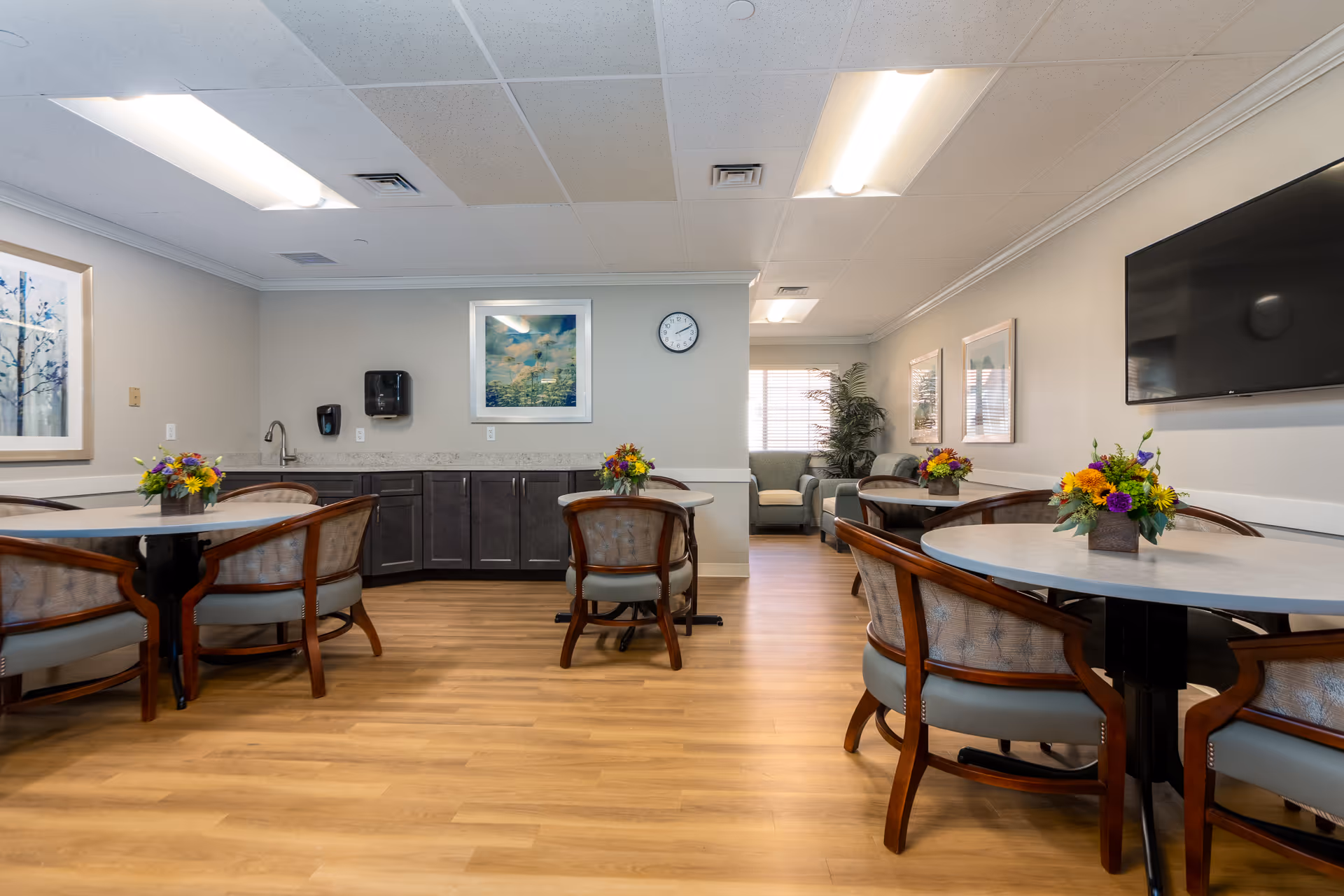 A well-lit common area with round tables and cushioned chairs arranged neatly. Each table has a small floral centerpiece. The room features wood flooring, light-colored walls with framed artwork, a wall-mounted TV, and a clock. In the background, there is a seating area with armchairs and a large window with blinds.