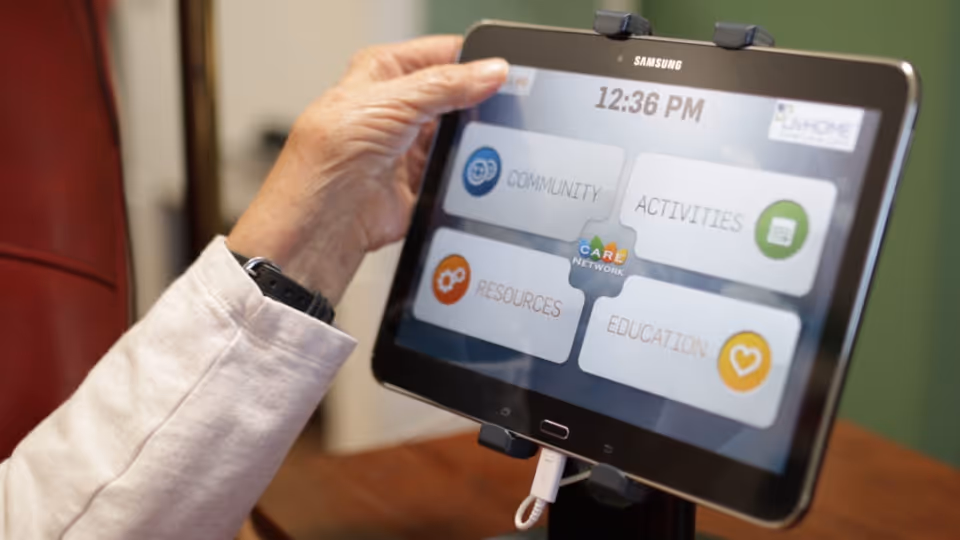 A hand holds a Samsung tablet displaying a senior living app with community and activities options.