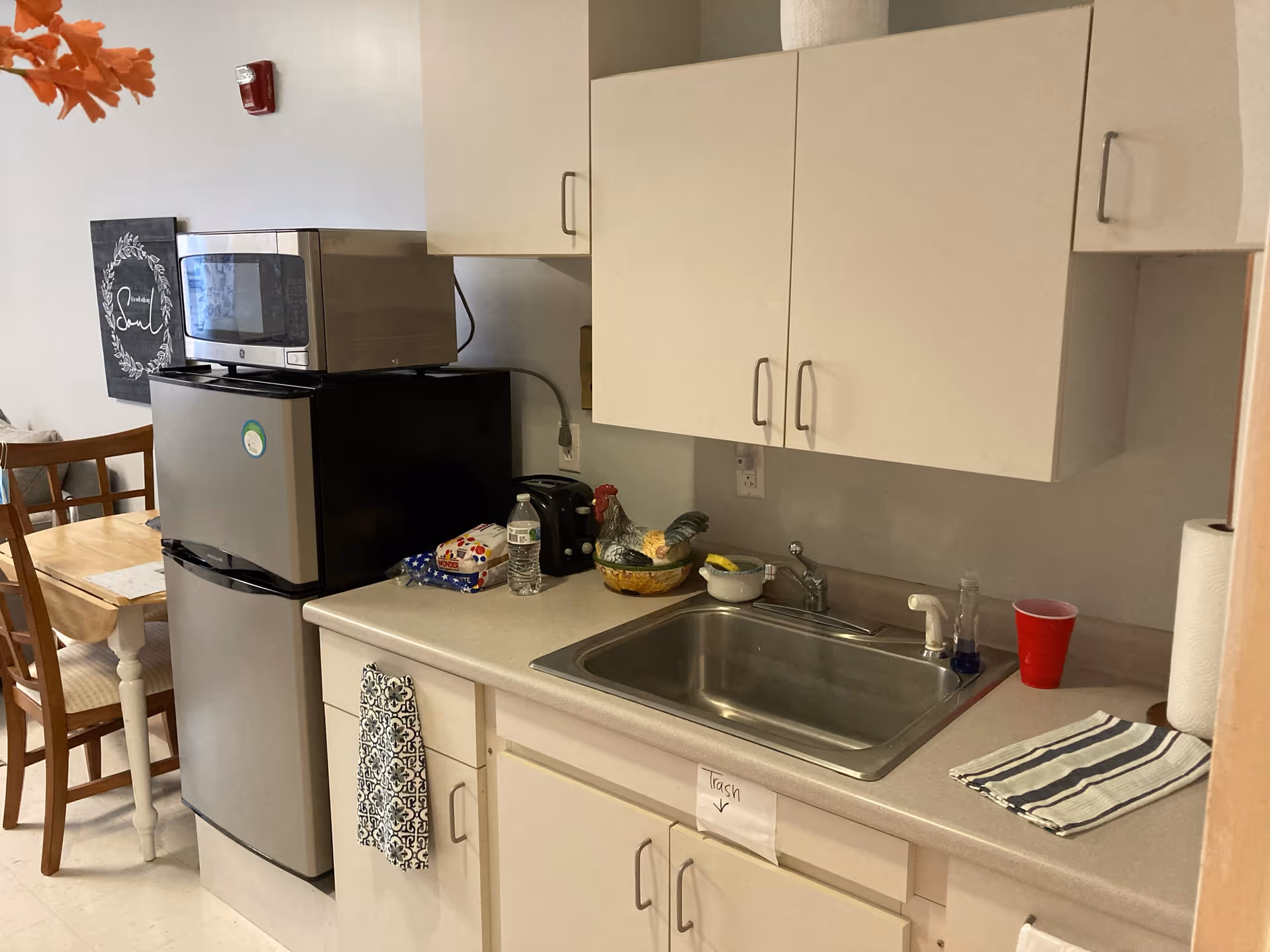 A small kitchen area with a stainless steel sink, white cabinets above and below the counter, a microwave on top of a mini refrigerator, a toaster, a loaf of bread, a water bottle, a decorative rooster bowl, a red plastic cup, paper towels, and a striped dish towel. In the background, there is a wooden dining table with chairs and a wall decoration.