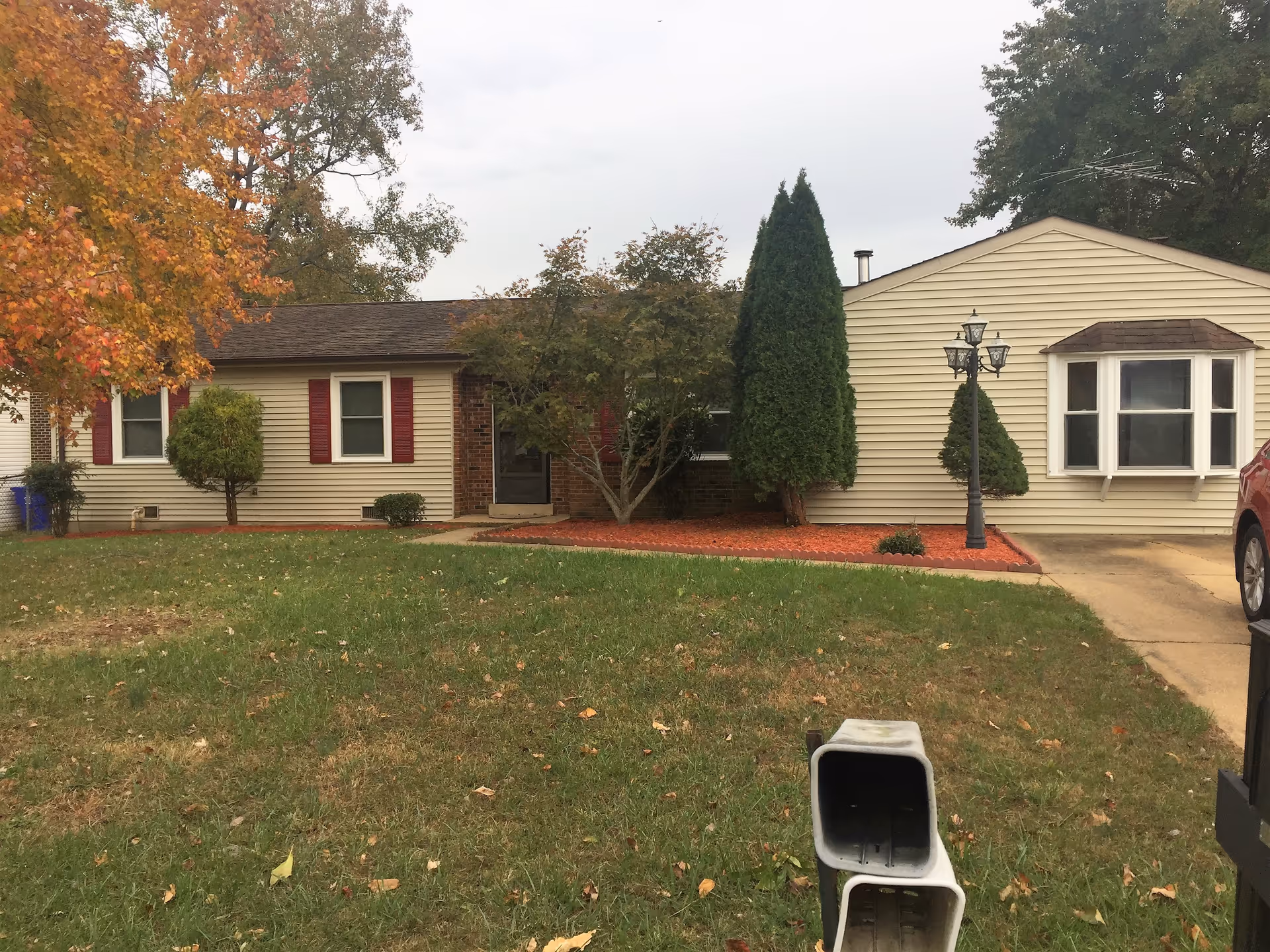 Single-story ranch-style house exterior with a front lawn, shrubs, a lamp post and driveway under an overcast sky.