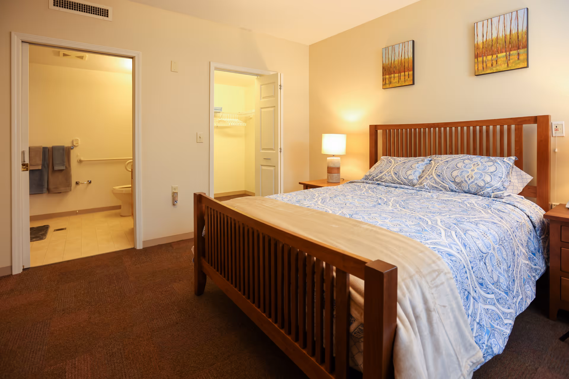 A cozy bedroom in a senior living facility with a wooden bed frame, blue patterned bedding, a bedside table with a lamp, and two paintings of trees on the wall. The room has an open door leading to a bathroom with towels hanging and a toilet visible, and another door leading to a walk-in closet with shelves and hanging space.