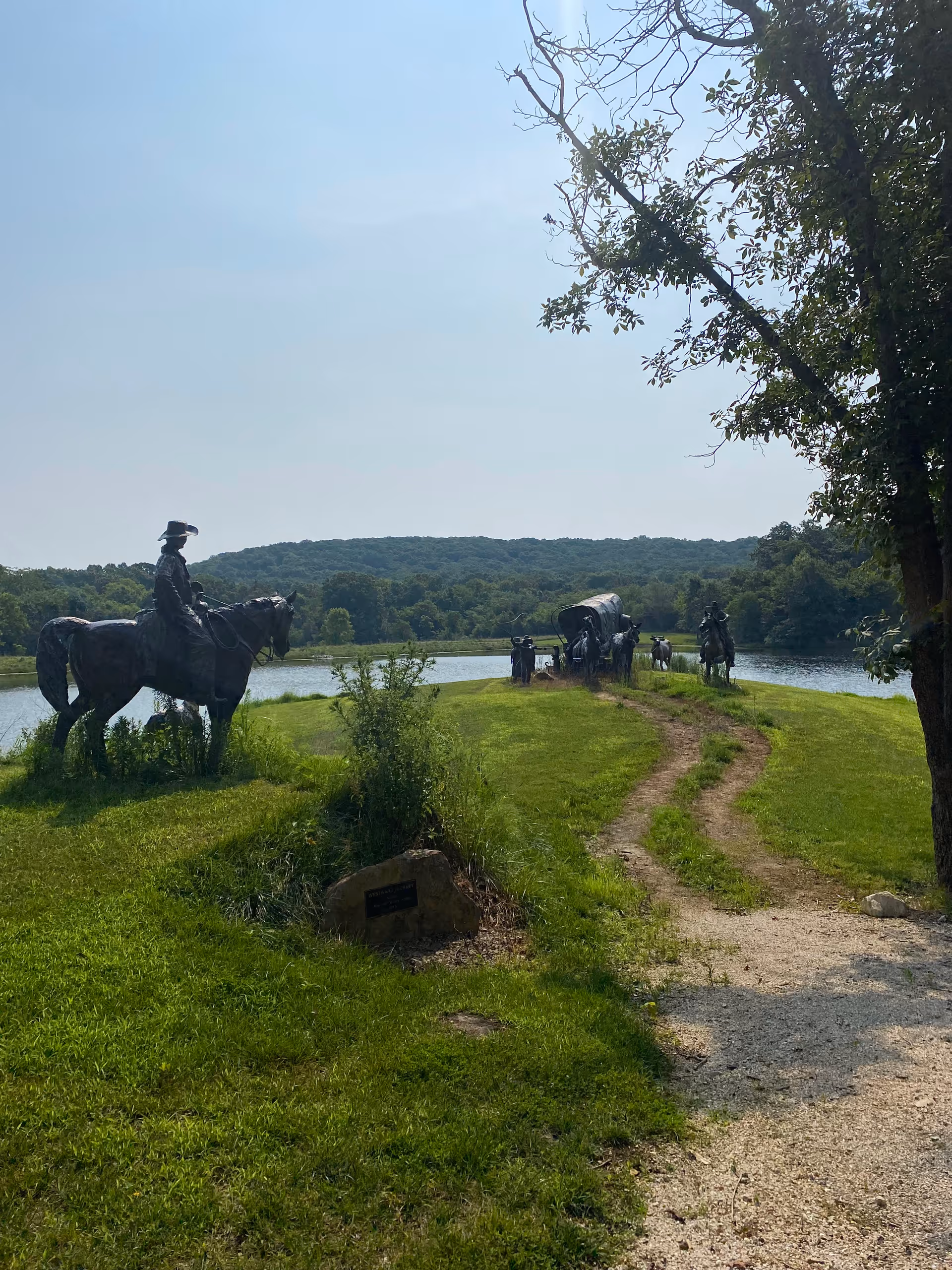 Outdoor scene of horse-mounted statues along a grassy path beside a lake.