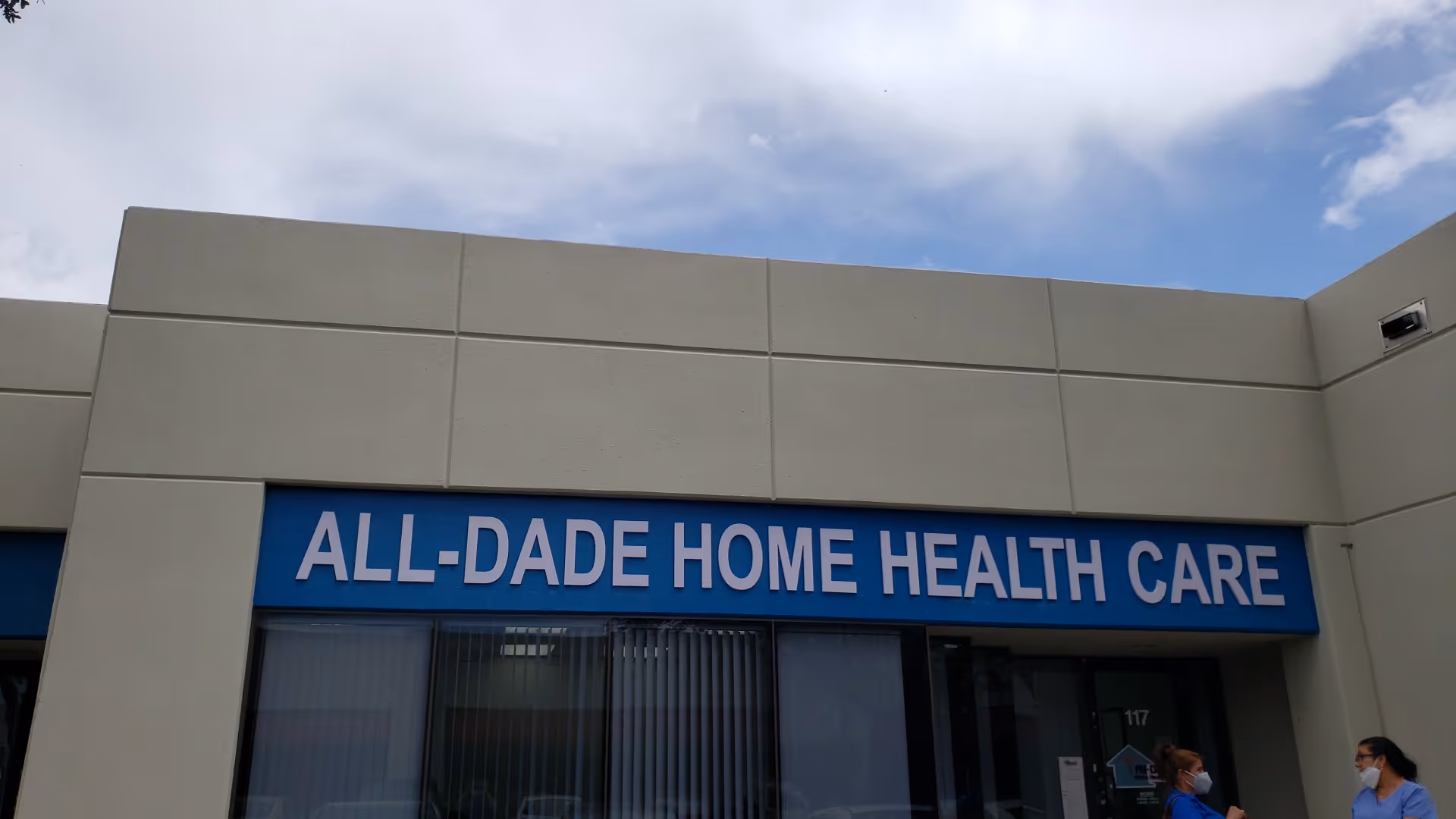 Exterior of All-Dade Home Health Care building with blue sign and two masked individuals
