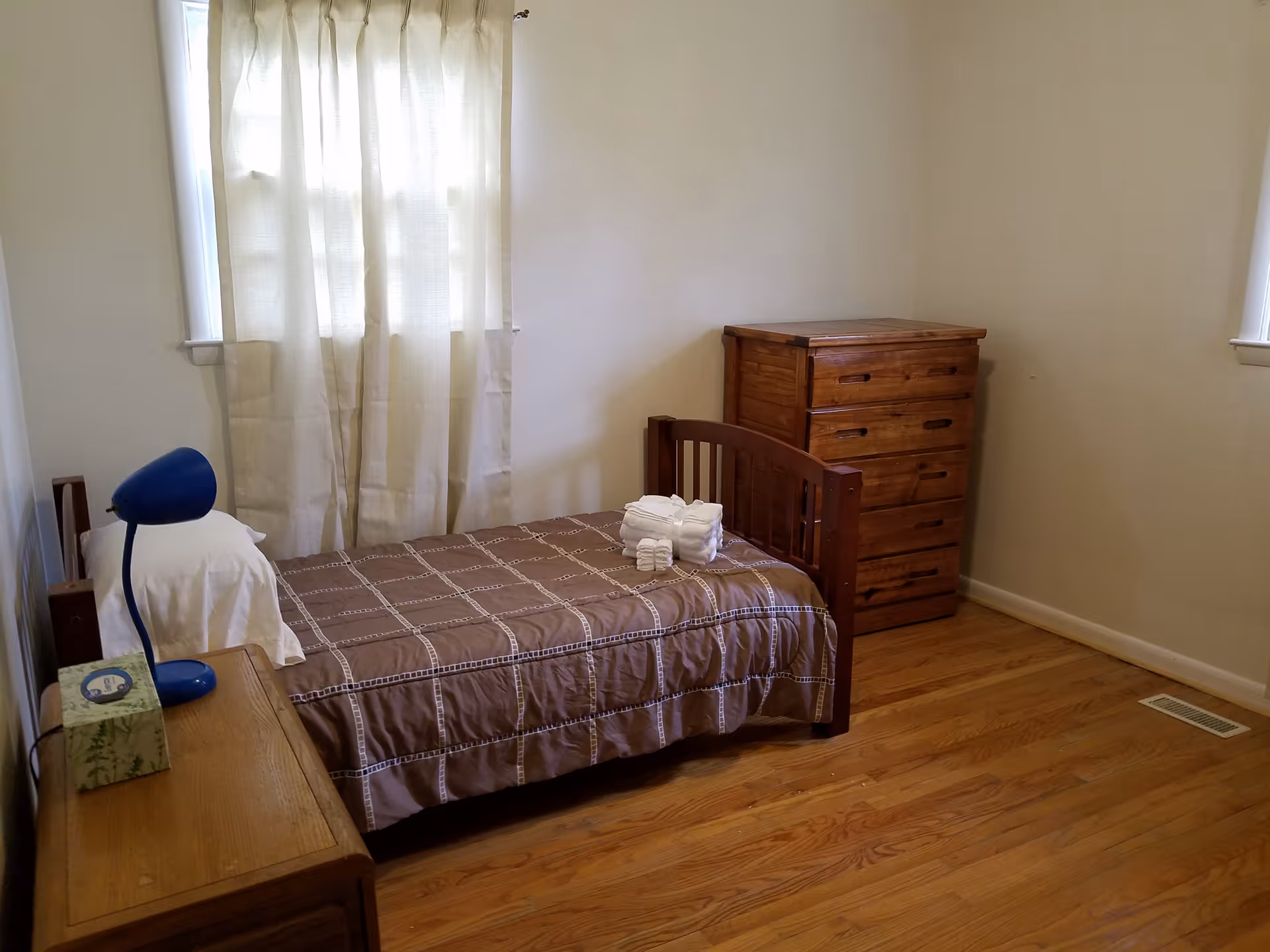 A simply furnished bedroom with single bed, dresser, nightstand, and curtained windows.