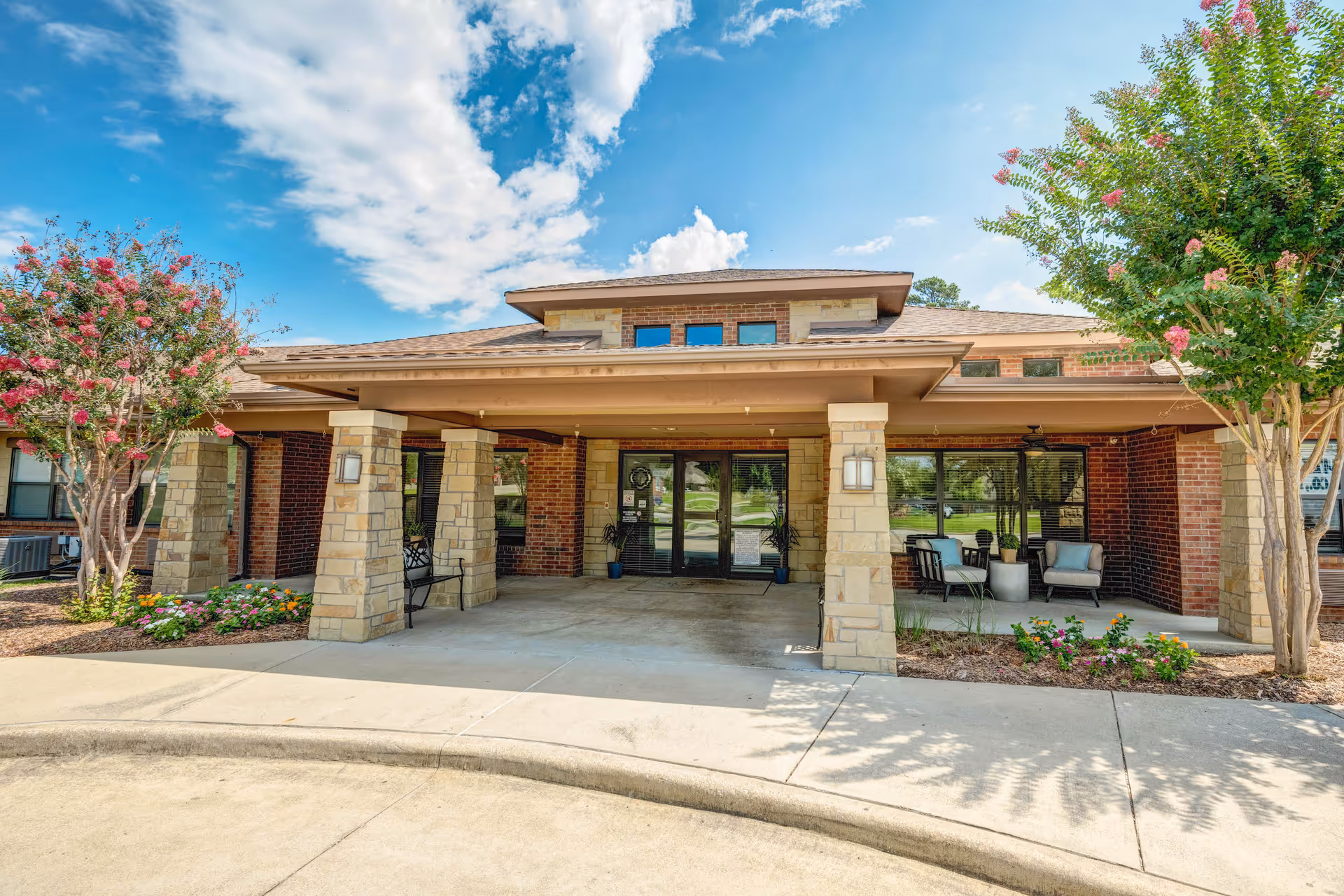 Front entrance of a single-story brick memory care building with a covered porte-cochere, stone columns, outdoor seating, and landscaped beds under a blue sky.