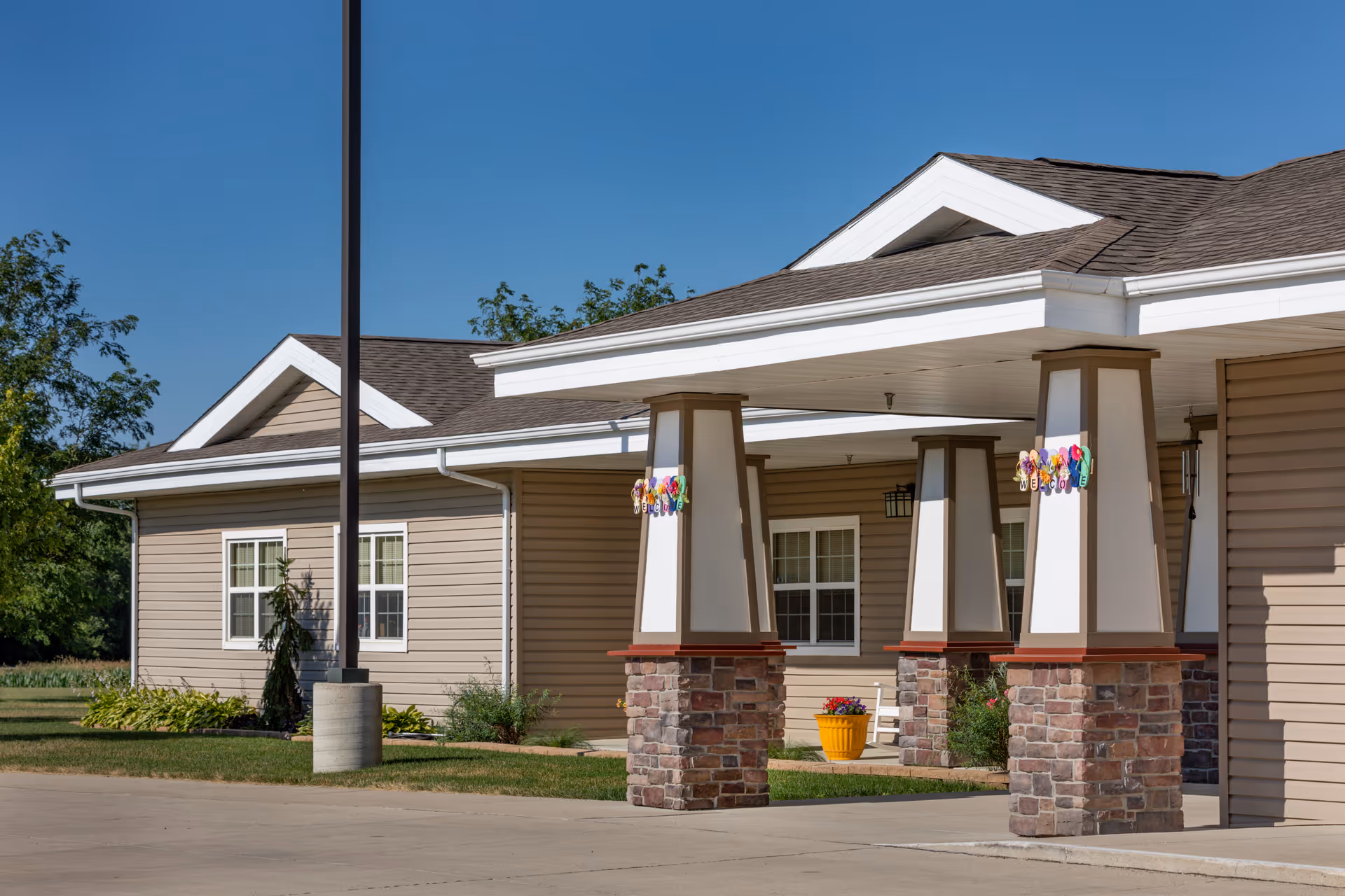 Front exterior of a beige assisted living building showing a covered entry portico with stone-clad columns, windows, and a potted plant.