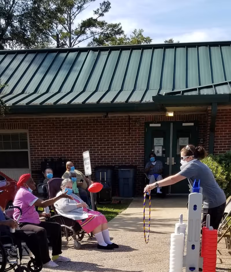 A group of elderly people sitting outside a brick building with a green metal roof, wearing masks. One elderly woman is tossing a red ring towards a staff member who is holding a purple and yellow ring. The staff member is also wearing a mask and appears to be facilitating an outdoor ring toss game. There are chairs and some trash bins near the building entrance.