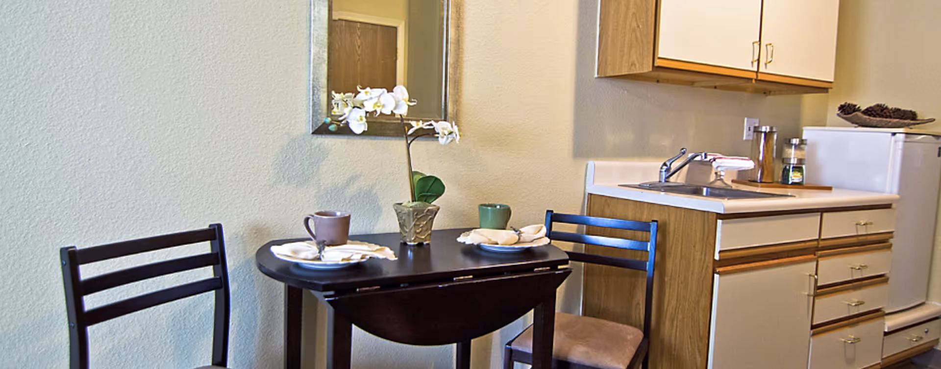Small dining table with two chairs, place settings, and an orchid beside a compact kitchen.