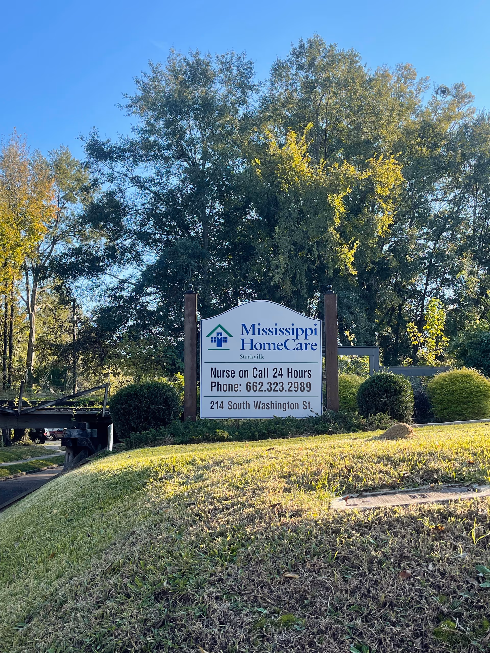 Outdoor scene featuring a Mississippi HomeCare sign on a grassy, tree-lined roadside.