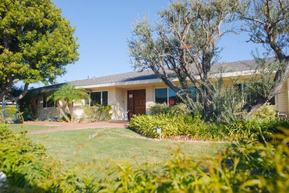 Single-story residential building with a front yard featuring green grass, various plants, and trees under a clear blue sky.