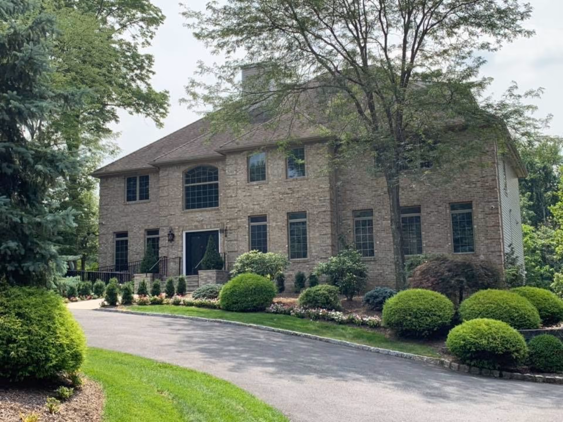 Two-story brick senior living building with manicured shrubs, trees, and a curved driveway in front.