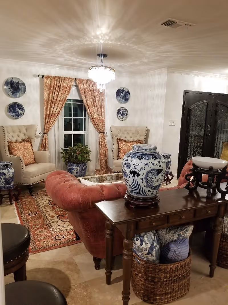 A warmly decorated living room with tufted chairs, drapery, plants, and blue-and-white porcelain vases.