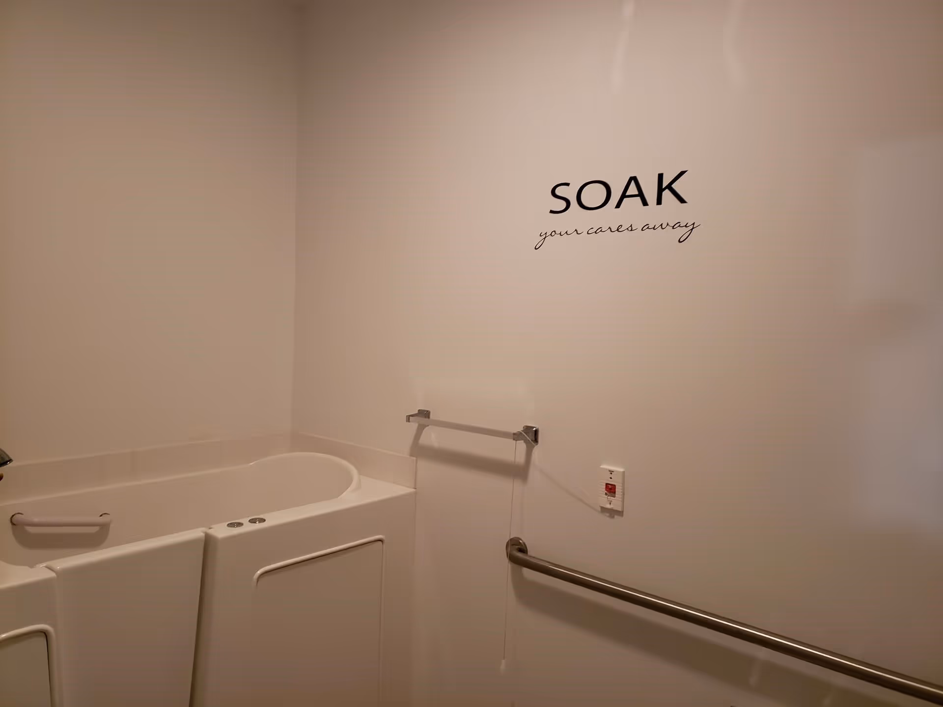 Interior bathroom with a walk-in bathtub, grab bars, towel rack, and a wall decal reading "SOAK your cares away".
