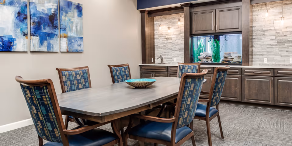 A dining room with a rectangular wooden table surrounded by six wooden chairs with blue patterned upholstery. On the table is a decorative bowl. The room features a wall with dark wooden cabinets and a built-in aquarium with green plants and rocks. Above the cabinets, there are two pendant lights and a textured stone backsplash. On the adjacent wall, there are three abstract blue and white paintings.