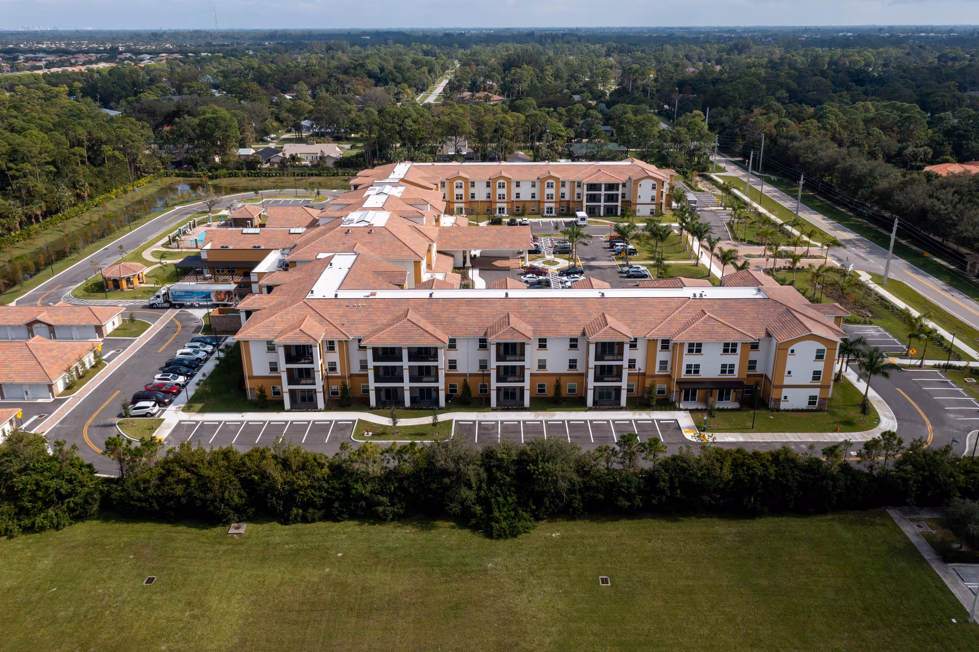 Aerial view of Palomino Gardens Retirement Community showing multiple connected buildings with red-tiled roofs, parking lots, surrounding greenery, and roads.