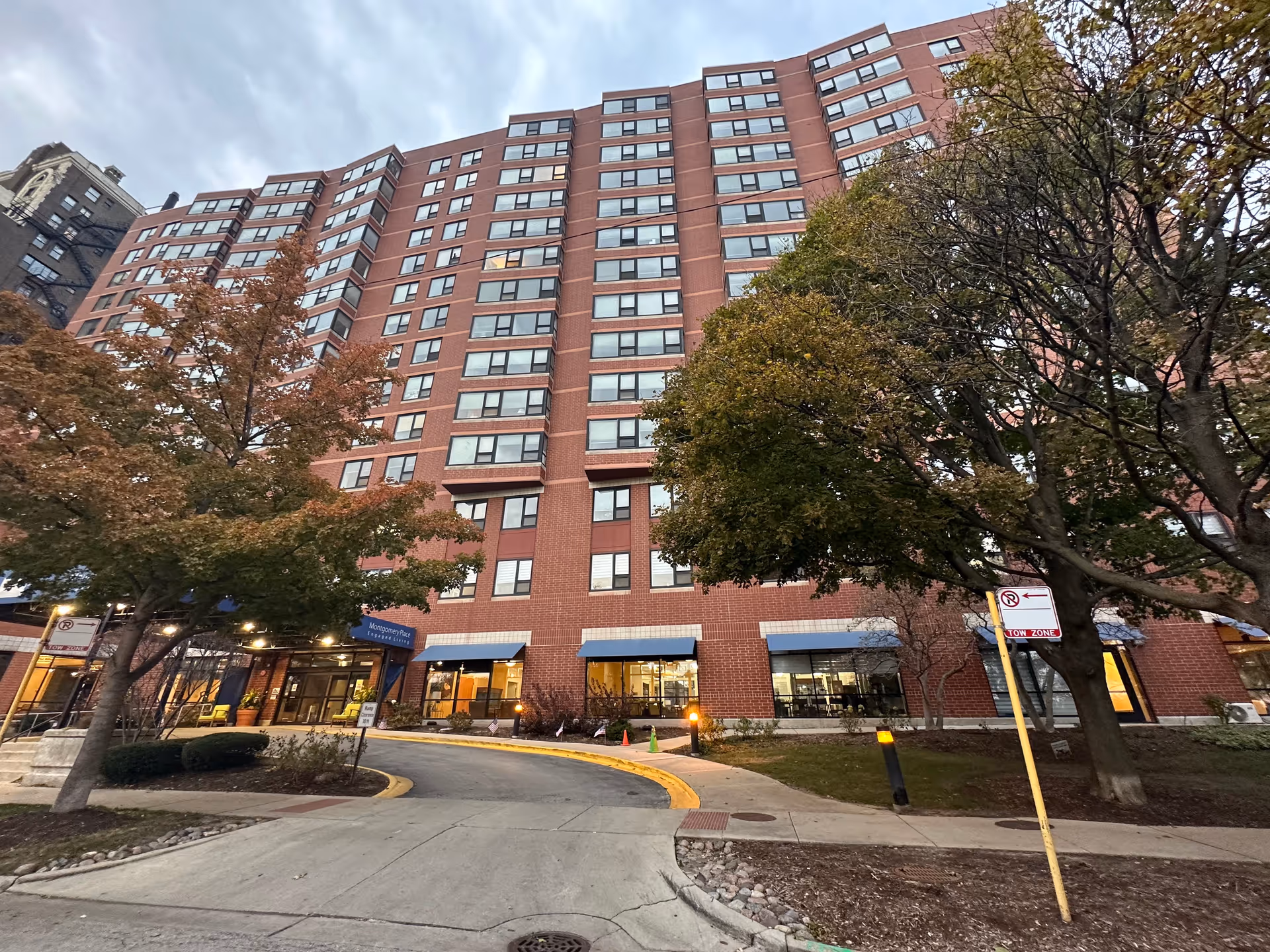 Exterior view of a multi-story brick building with many windows, identified as Montgomery Place Retirement Community. The entrance is visible with a blue awning and surrounded by trees with autumn foliage. The sky is cloudy and the driveway curves toward the entrance.