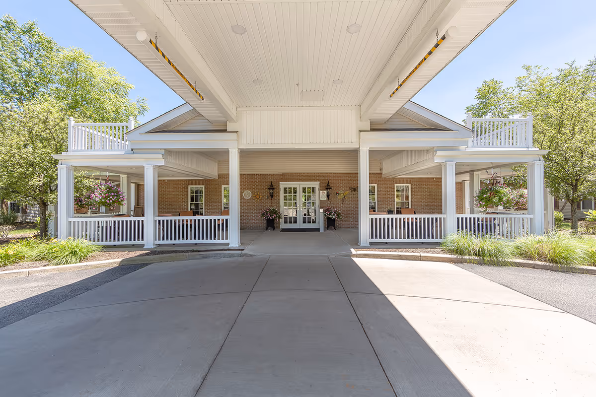 Covered entrance and porte-cochere of a brick assisted living building with white columns, railings, and hanging flower baskets.