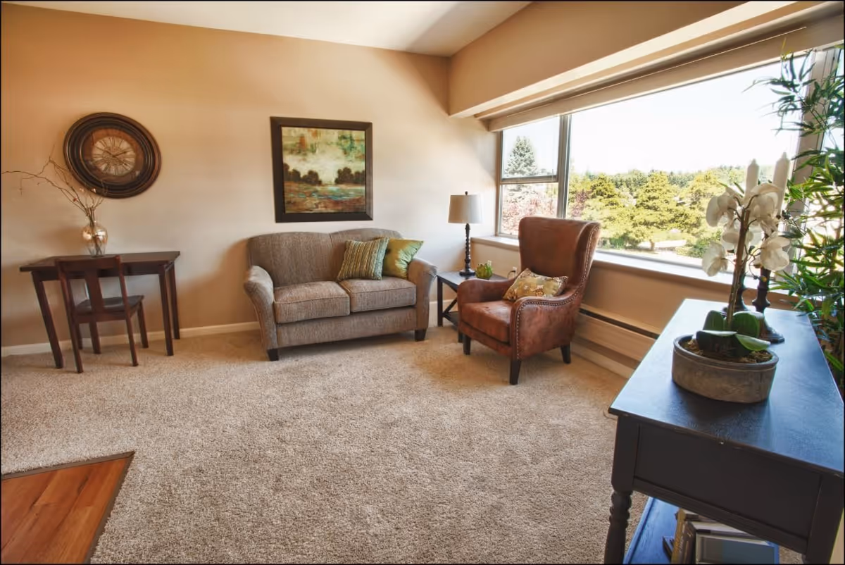 Cozy senior living lounge with sofa, armchair, tables, and a large window view.