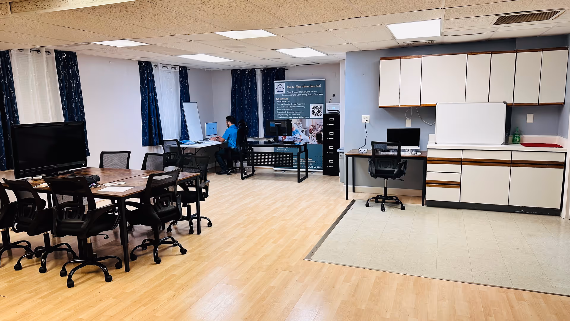 Interior communal room with tables, chairs, computers, and a kitchenette in a senior facility.