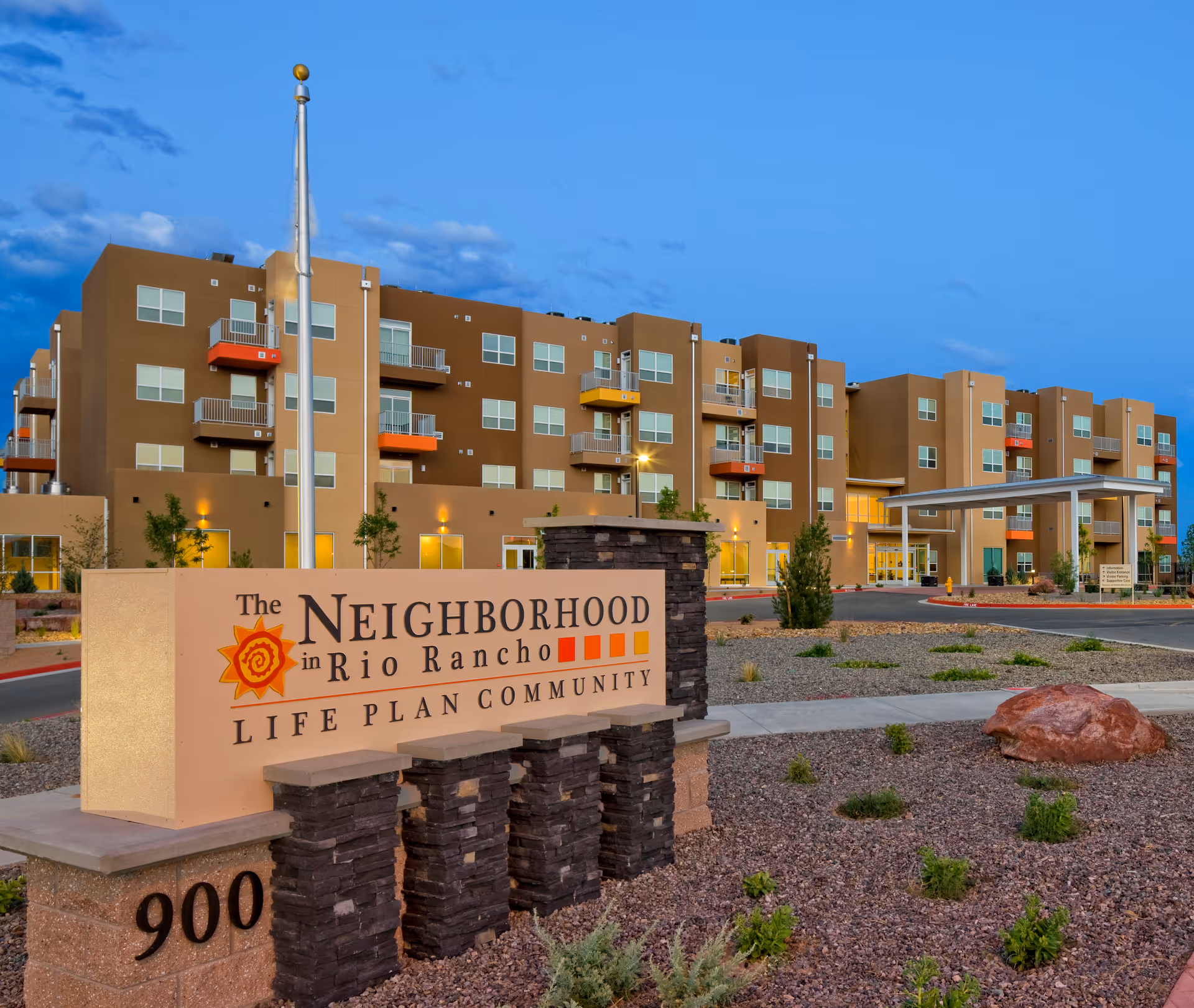 Exterior view of The Neighborhood in Rio Rancho Life Plan Community building during dusk with a clear blue sky. The multi-story building has balconies with colorful railings and a covered entrance. In the foreground, there is a large sign with the community name and address 900, surrounded by desert landscaping with rocks and small plants.