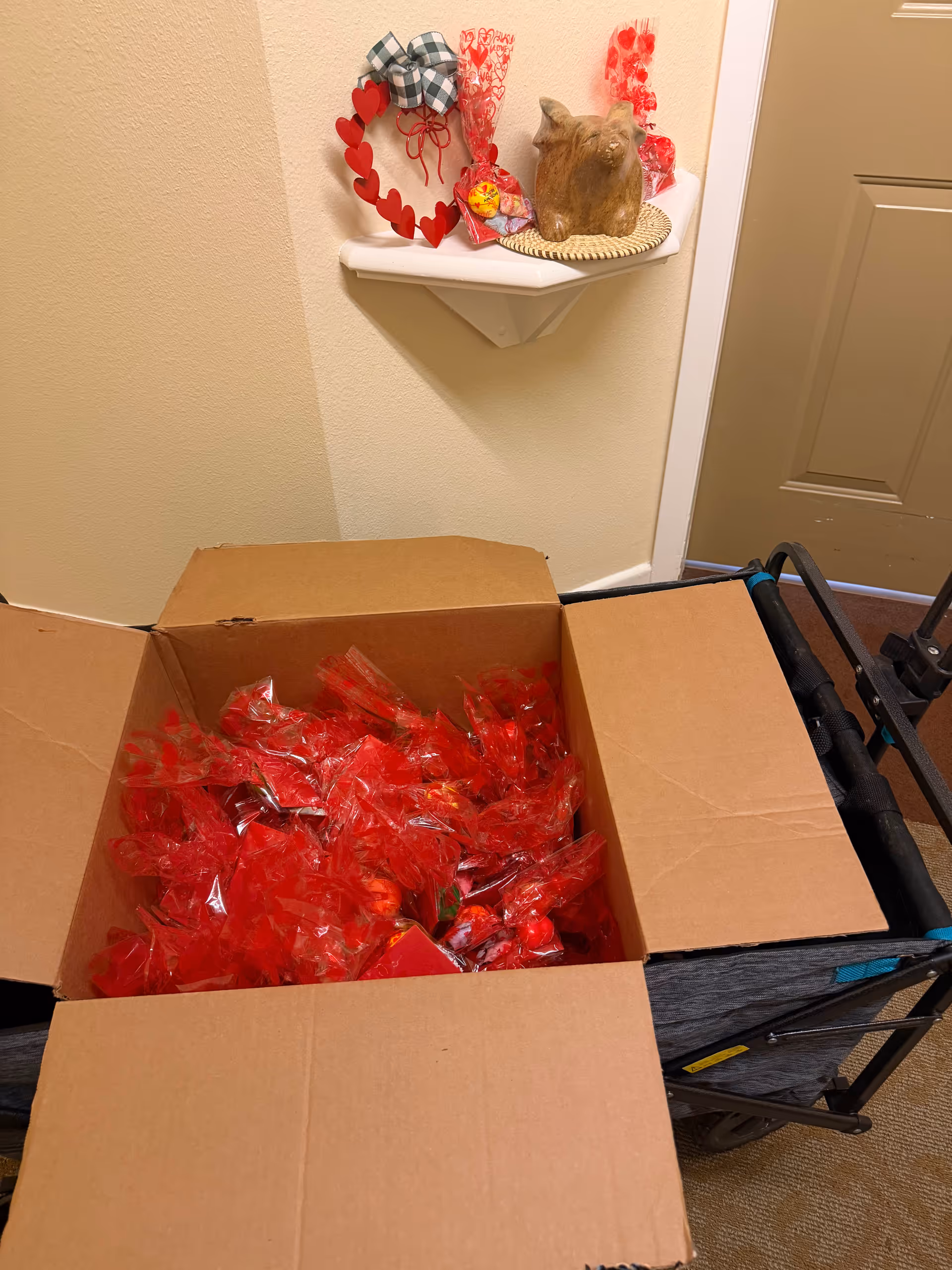 Cardboard box filled with red-wrapped items on a cart in a hallway near Valentine decor.