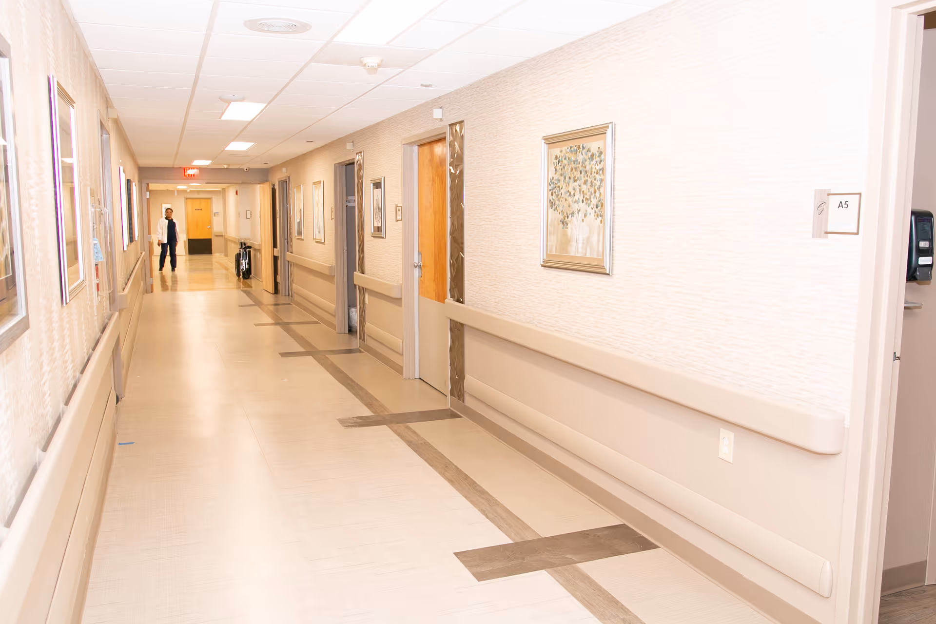 A long, well-lit healthcare hallway with handrails, framed artwork, several doors and a person at the far end.