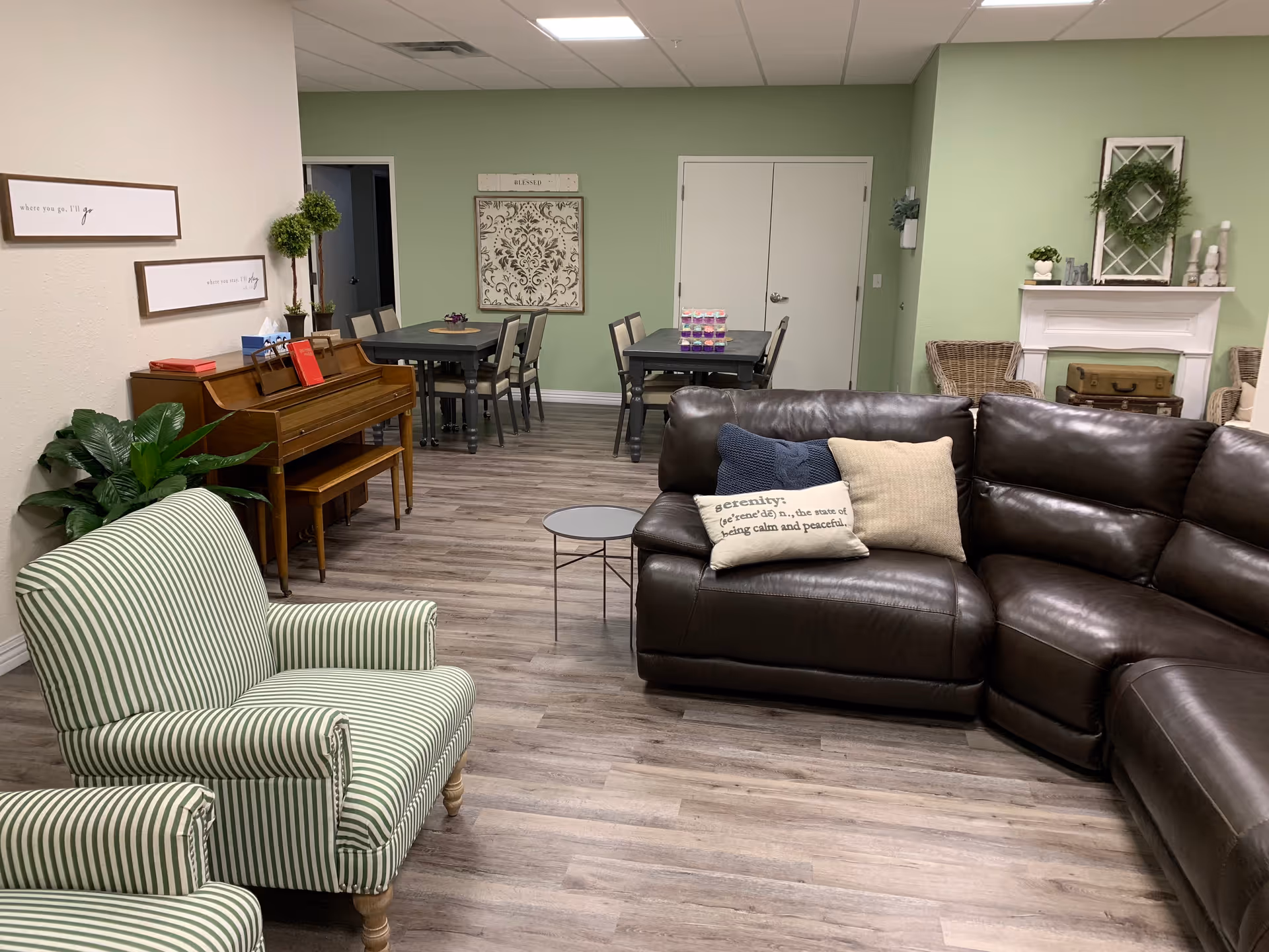 A cozy living room area with a dark brown leather sectional sofa adorned with decorative pillows, including one with the word 'serenity' on it. Two green and white striped armchairs are positioned nearby. In the background, there is a wooden piano with a bench, two tables with chairs, and a white fireplace decorated with a green wreath and small plants. The walls are painted light green and beige, and the floor is wood laminate.
