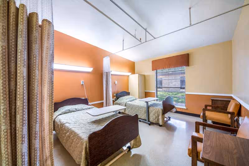 Two beds with beige bedspreads, privacy curtains, and seating in a shared patient room.