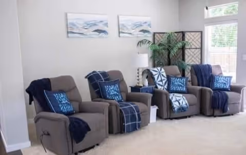 A cozy living room area with three gray recliner chairs, each adorned with blue and white patterned pillows and navy blue throws. Behind the chairs is a white wall with two abstract ocean-themed paintings. A white lamp and a green potted plant are placed between the chairs, and a folding screen is visible in the corner near a window with blinds.