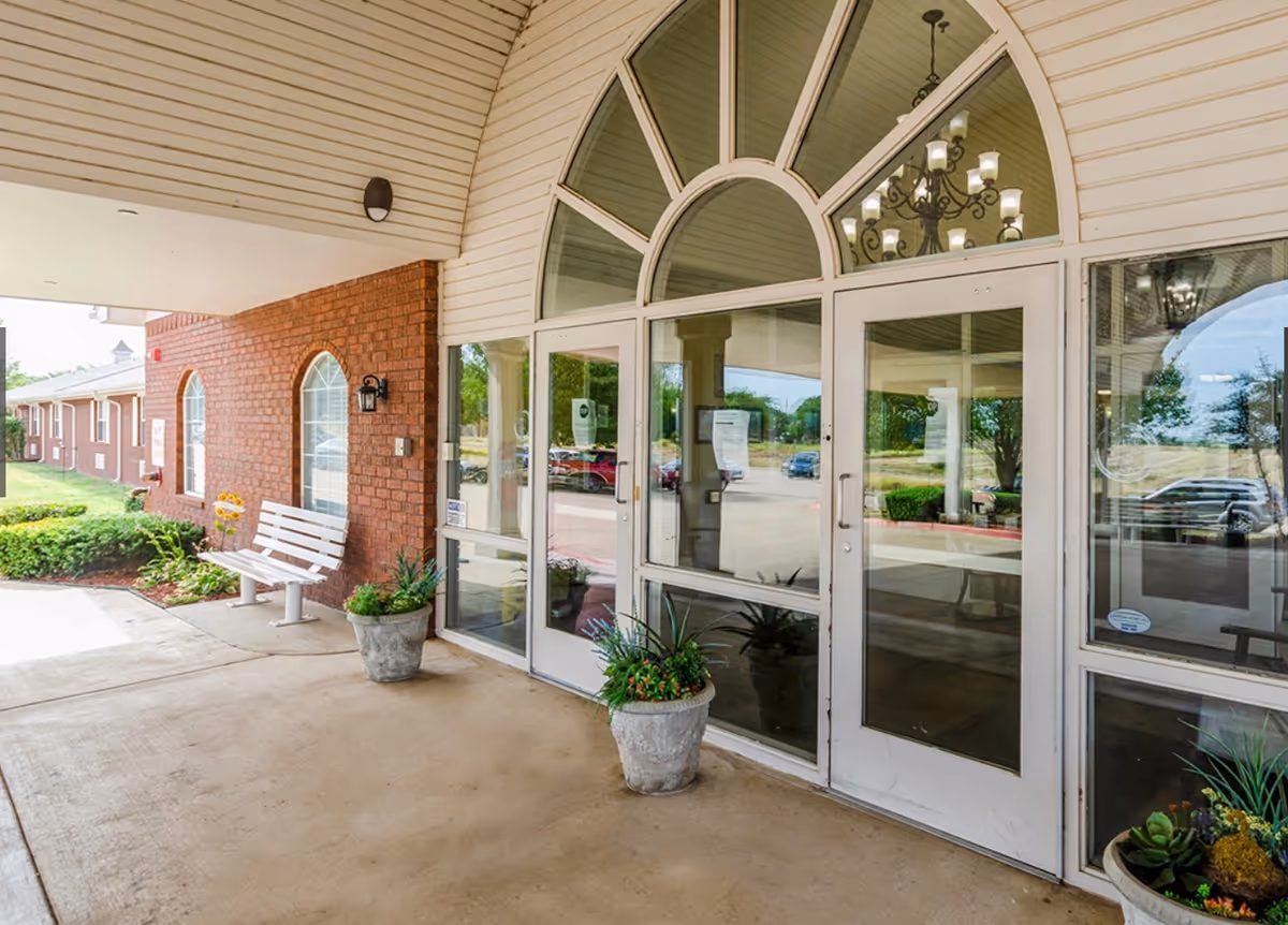 Covered front entrance with glass double doors and an arched transom, potted plants, and a white bench beside a brick wall.