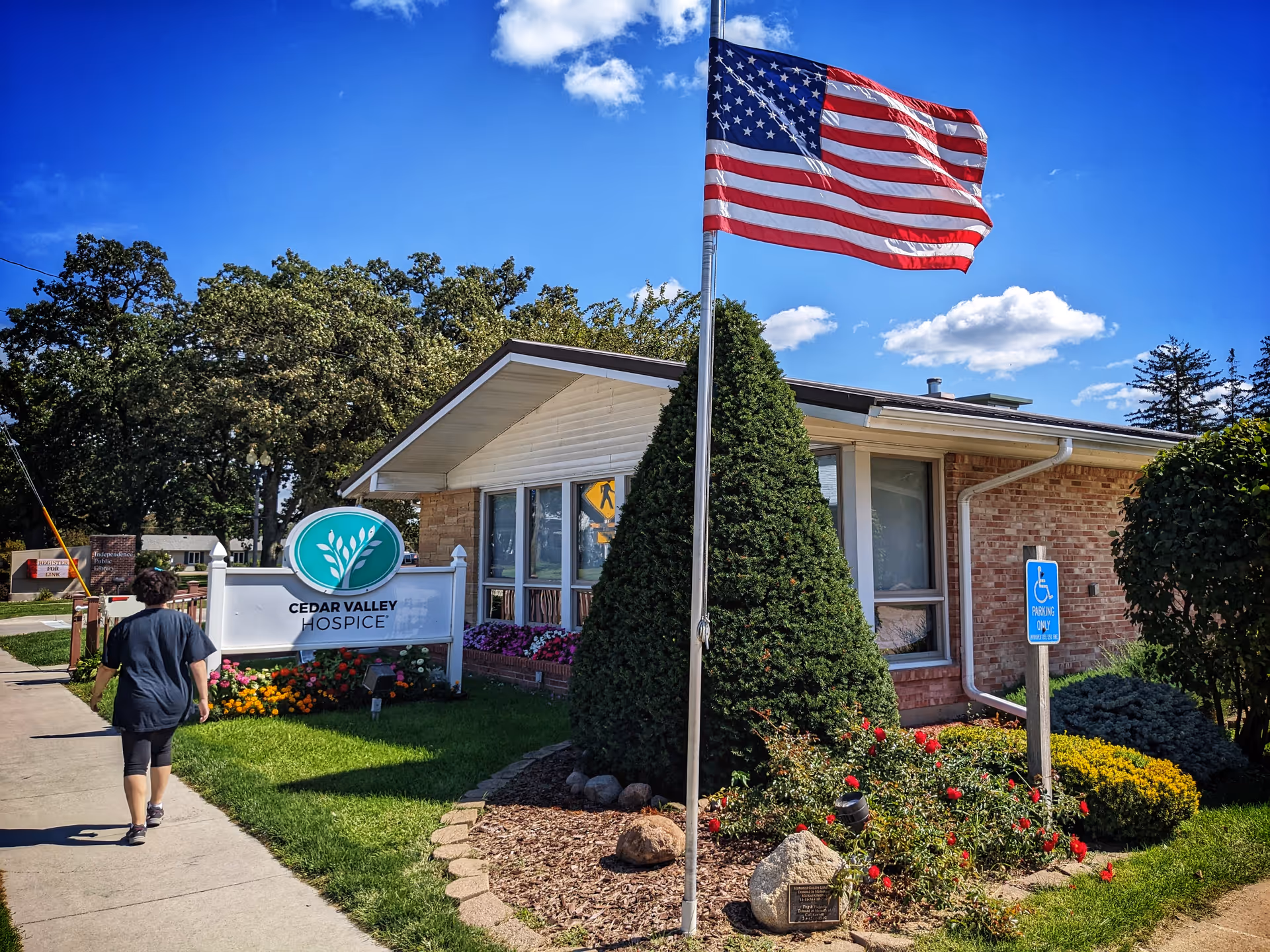 Exterior view of a Cedar Valley Hospice building with flag, landscaping, and a person walking.