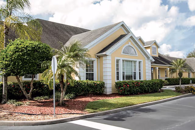Sunlit yellow single-story building with white trim, landscaped beds and palm trees along a driveway.