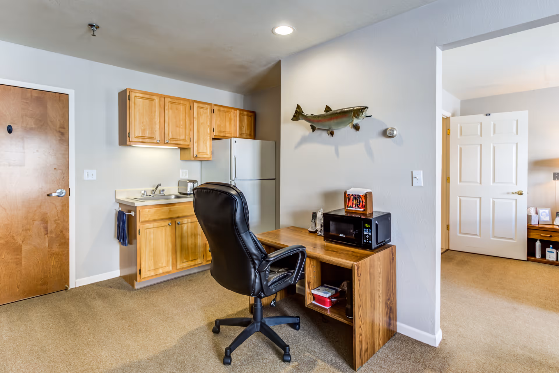 Compact kitchenette and desk area with wooden cabinets, fridge, sink, and microwave.