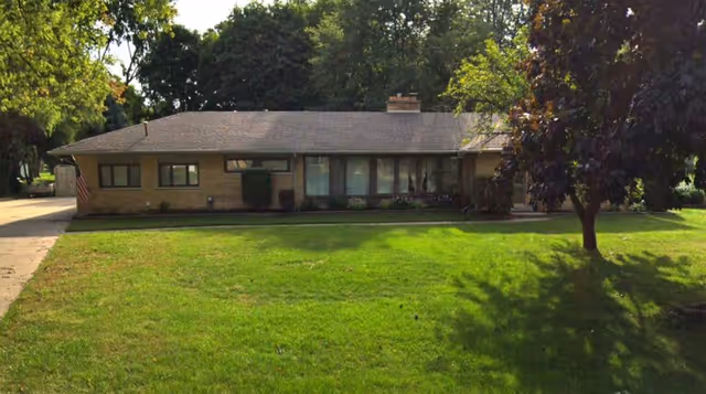 Single-story ranch-style house with a large front lawn, driveway, and trees.